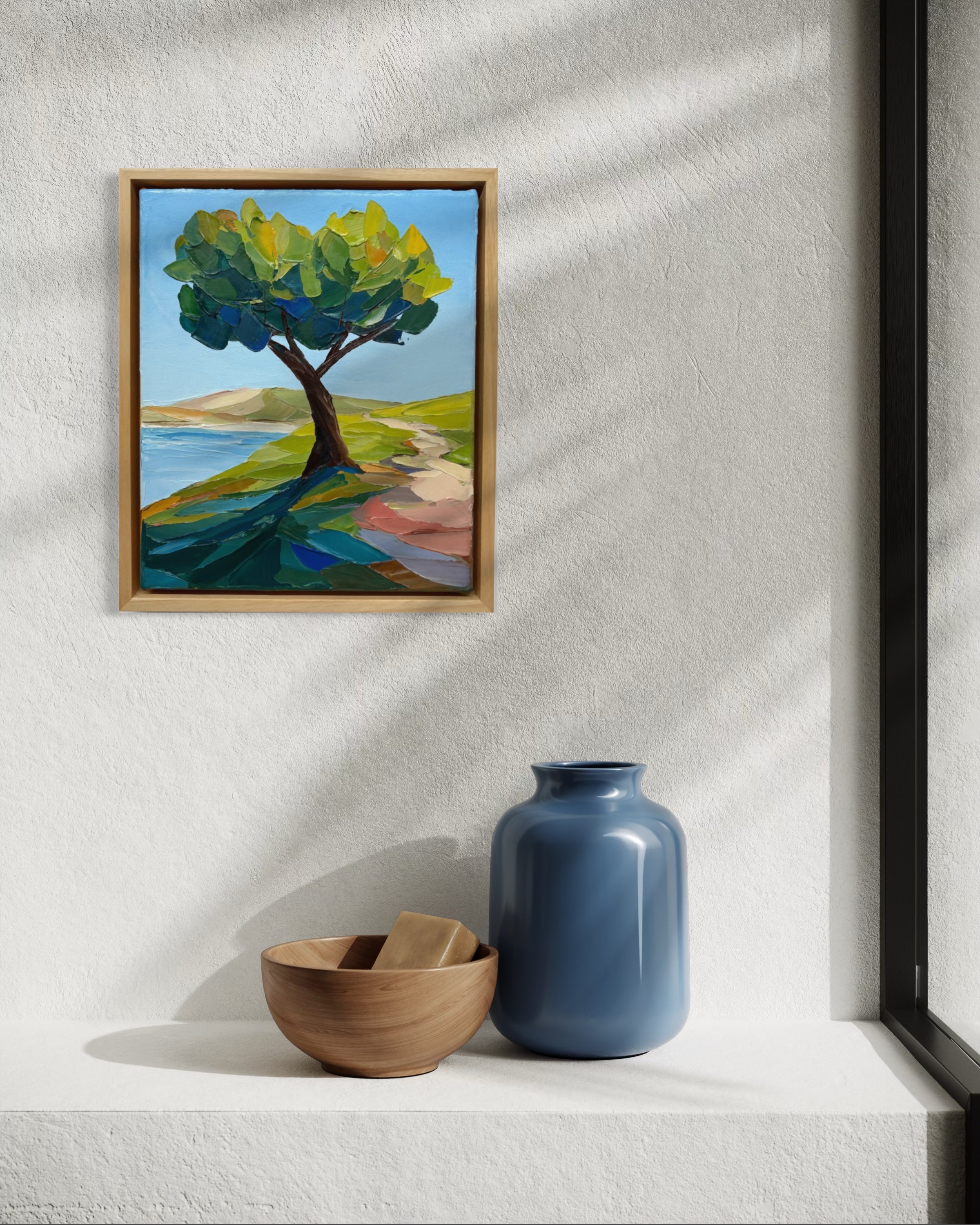 Framed painting of a tree on a wall with a blue vase and wooden bowl below.