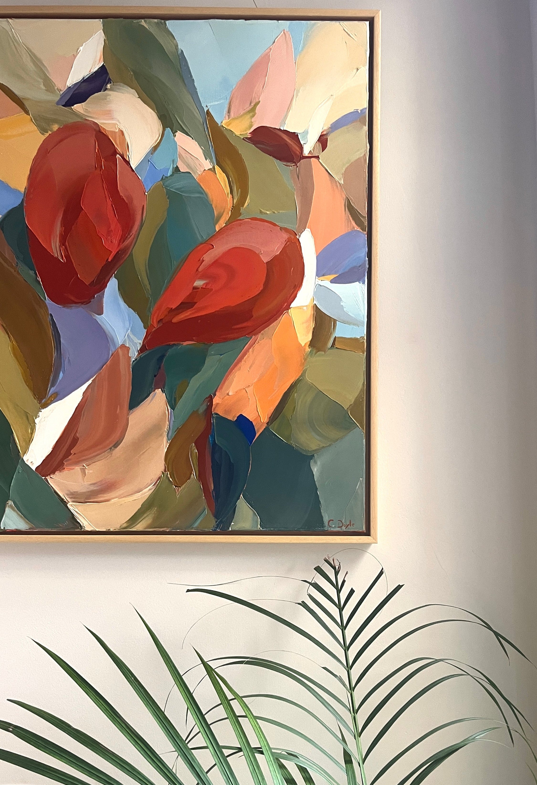 Abstract floral painting in a gold frame on a wall with green leaves in the foreground.