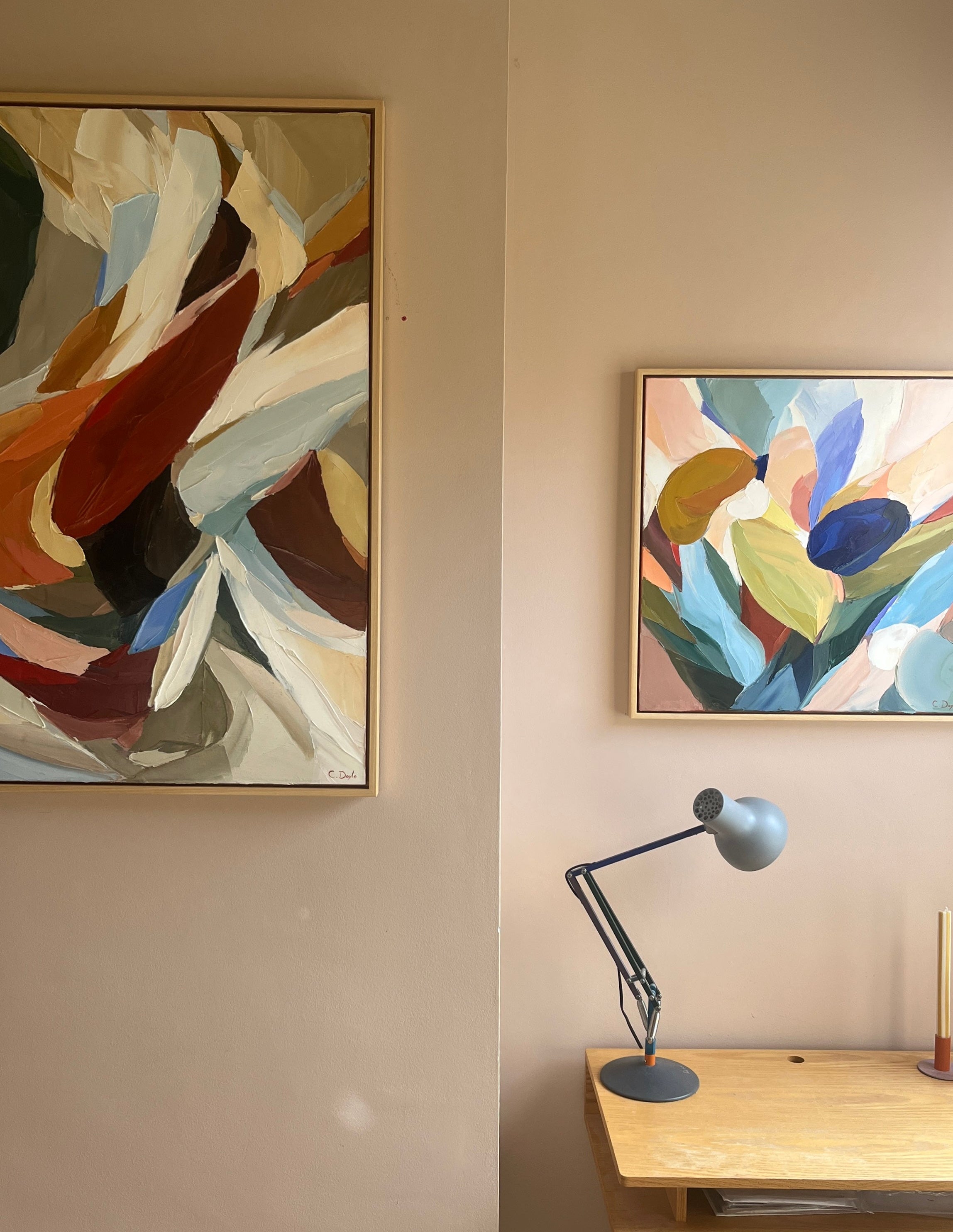 Two abstract paintings on a wall with a desk and lamp below.