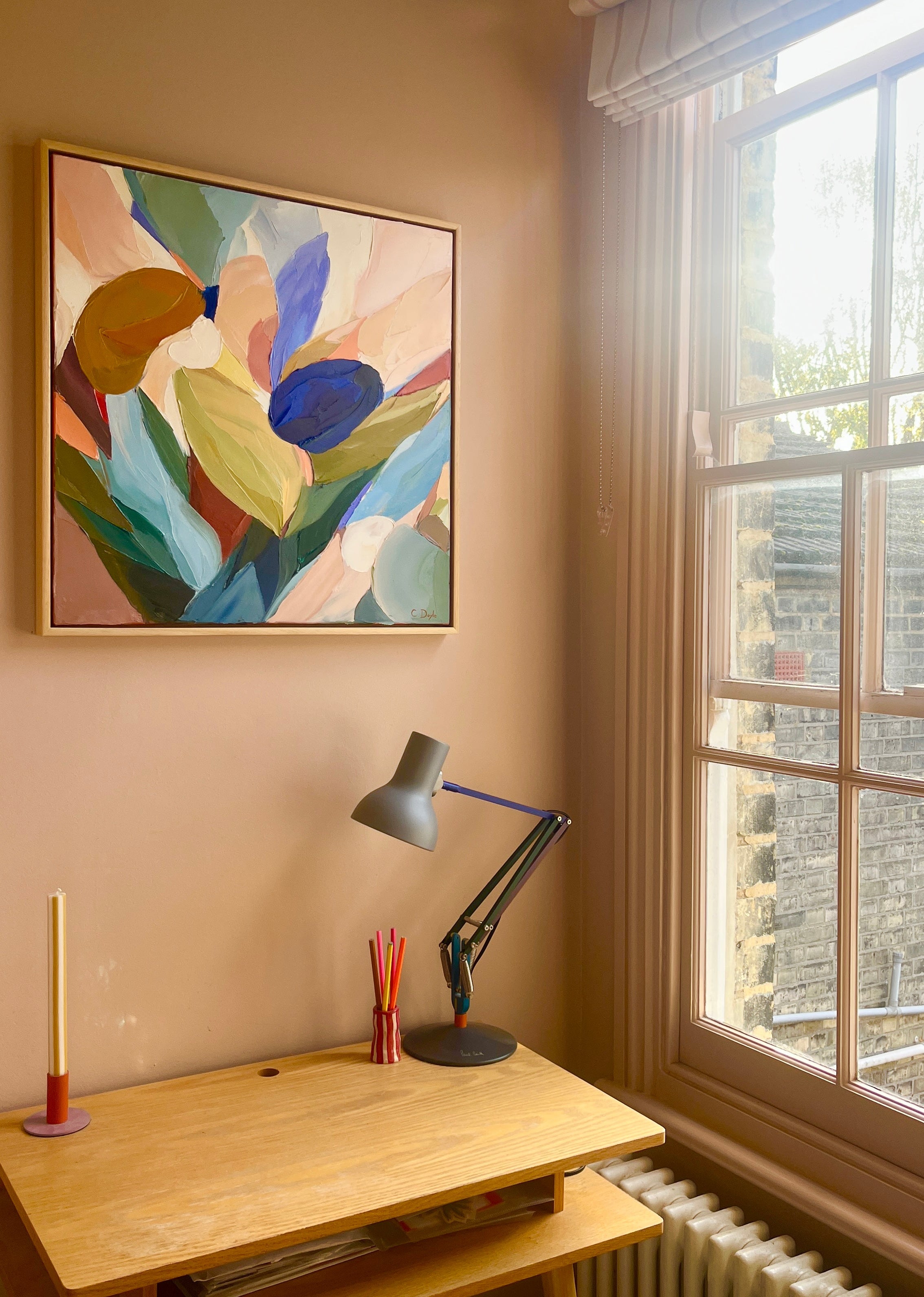 Desk with lamp, colorful abstract painting on wall, and window in room.