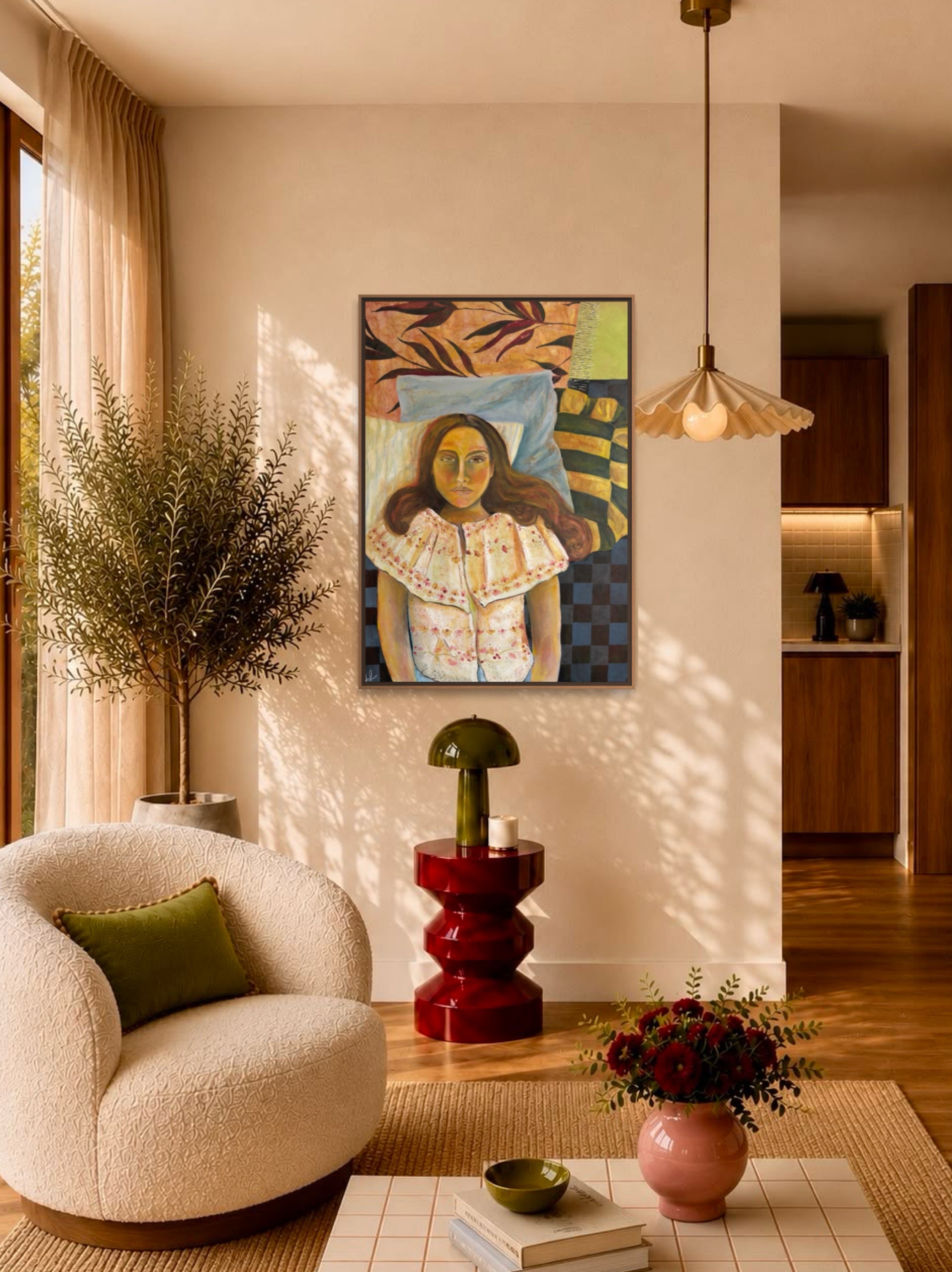 A framed art print featuring a stylized portrait of a woman hangs on a beige wall in a modern, sunlit living room with a cream boucle armchair, a red side table, and a hanging pendant light.