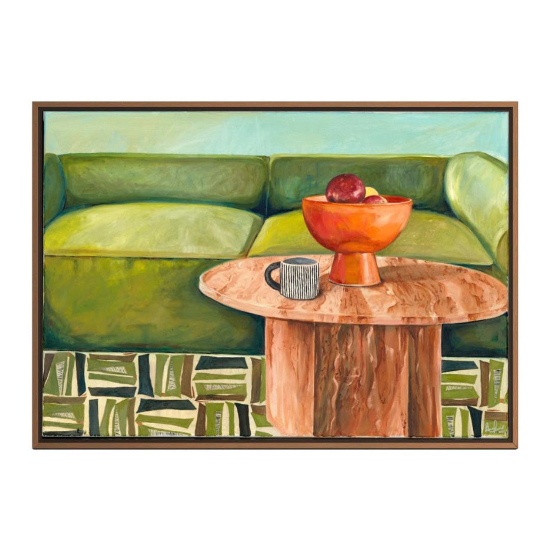 Framed artwork of a green sofa with a wooden table and orange bowl containing apples.