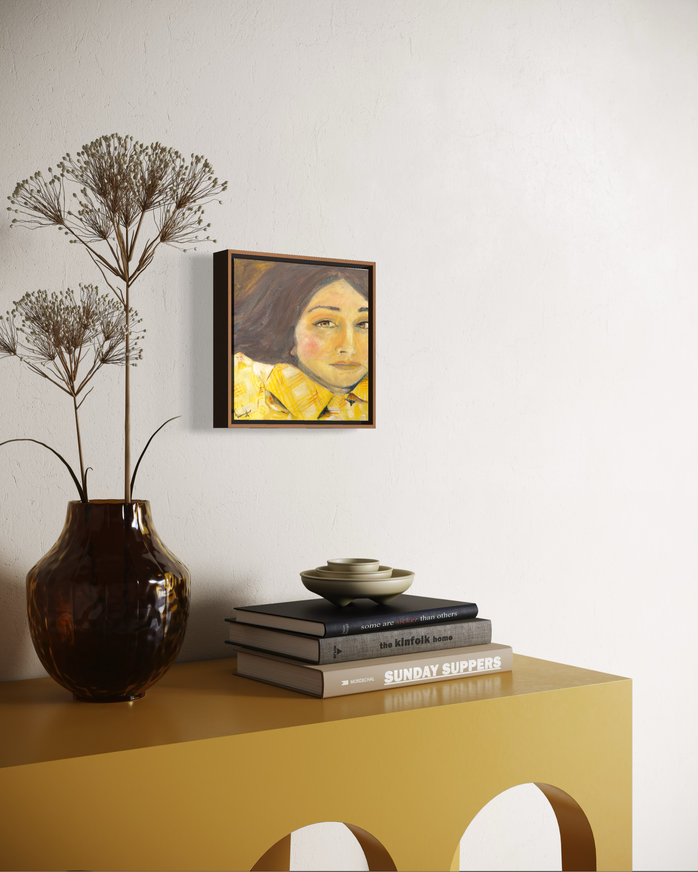 A framed portrait painting of a woman in a yellow top hangs on a white wall above a mustard yellow console table styled with a dark amber vase, dried flowers, and books.