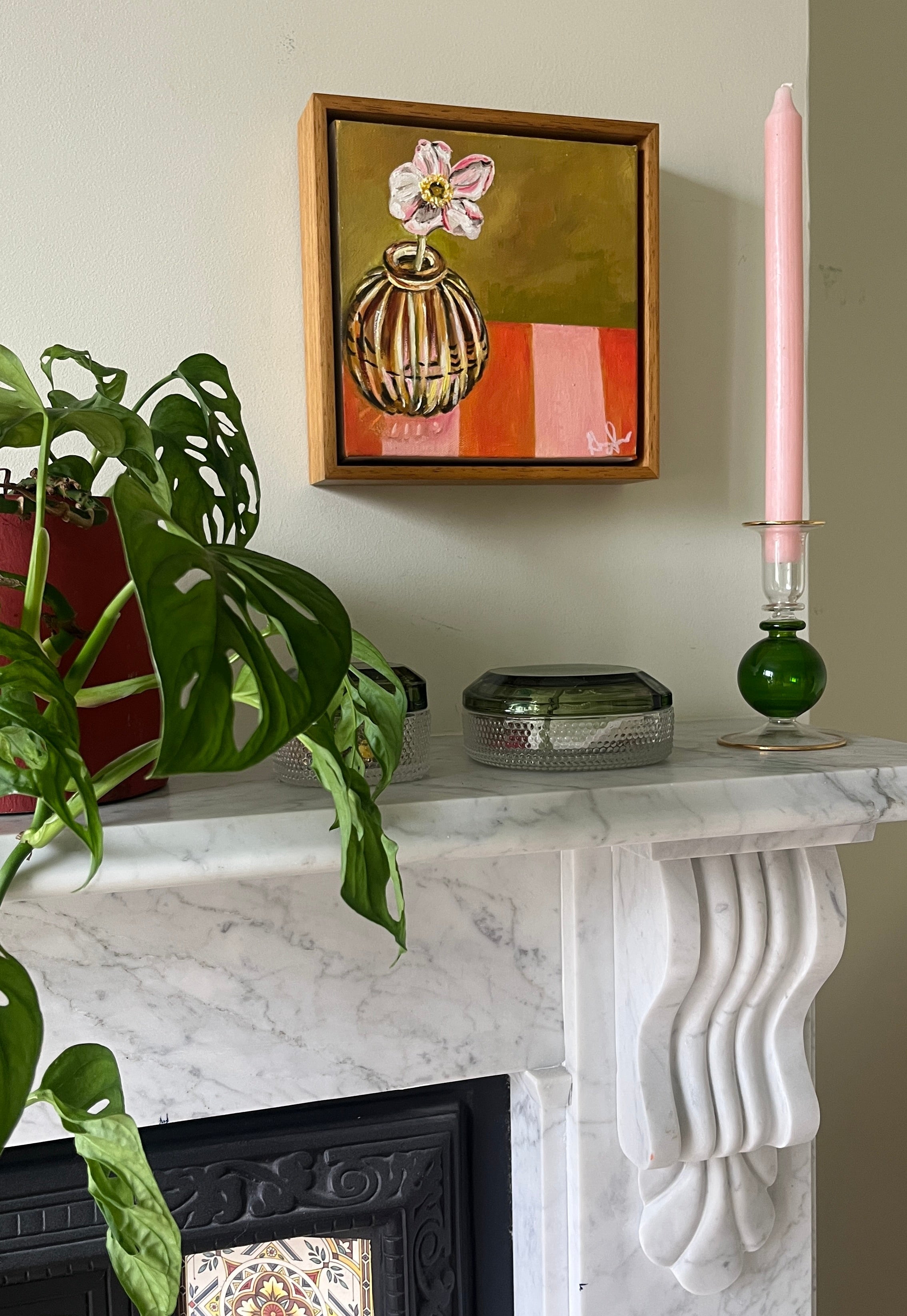 Framed artwork on a marble fireplace mantel with a plant and candle.