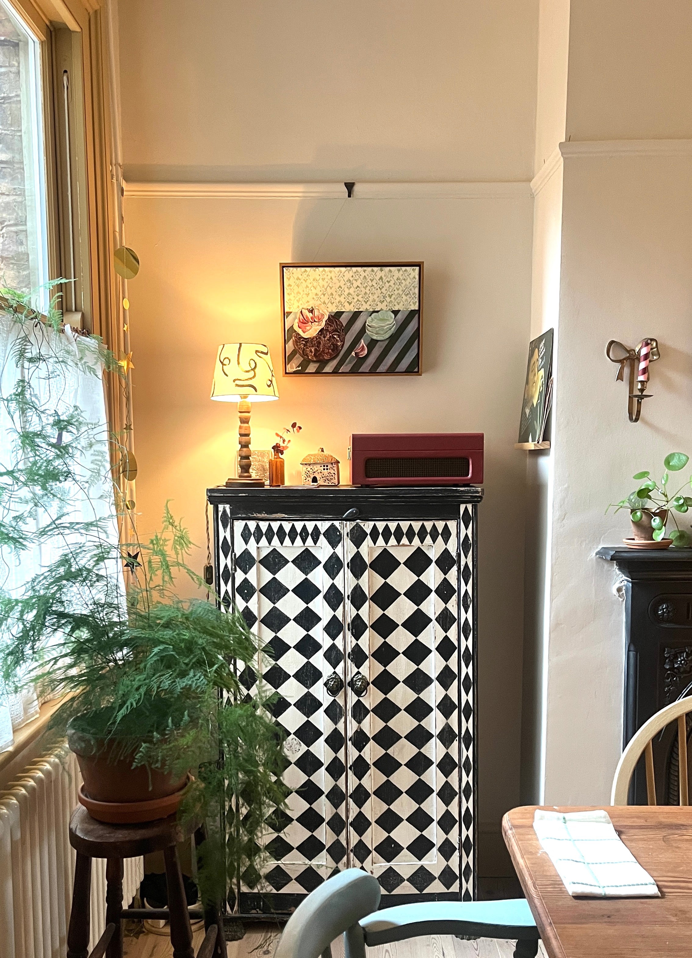 Narrow room with a black and white patterned cabinet, plants, and a table. Art work with oak frame with image of garlic and striped table cloth.