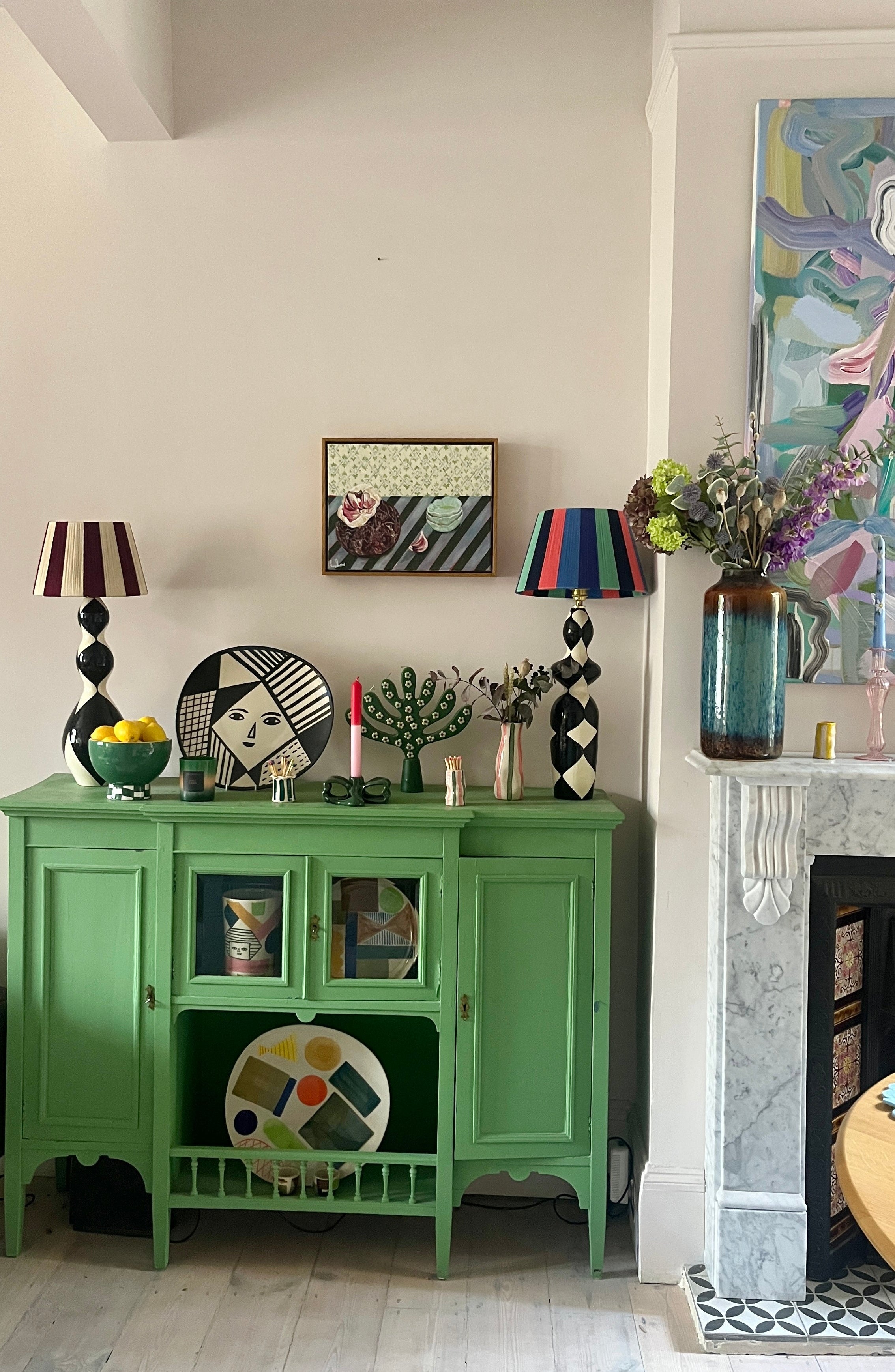 Green cabinet with decorative items in a room with a fireplace and colorful artwork and striped lampshades.