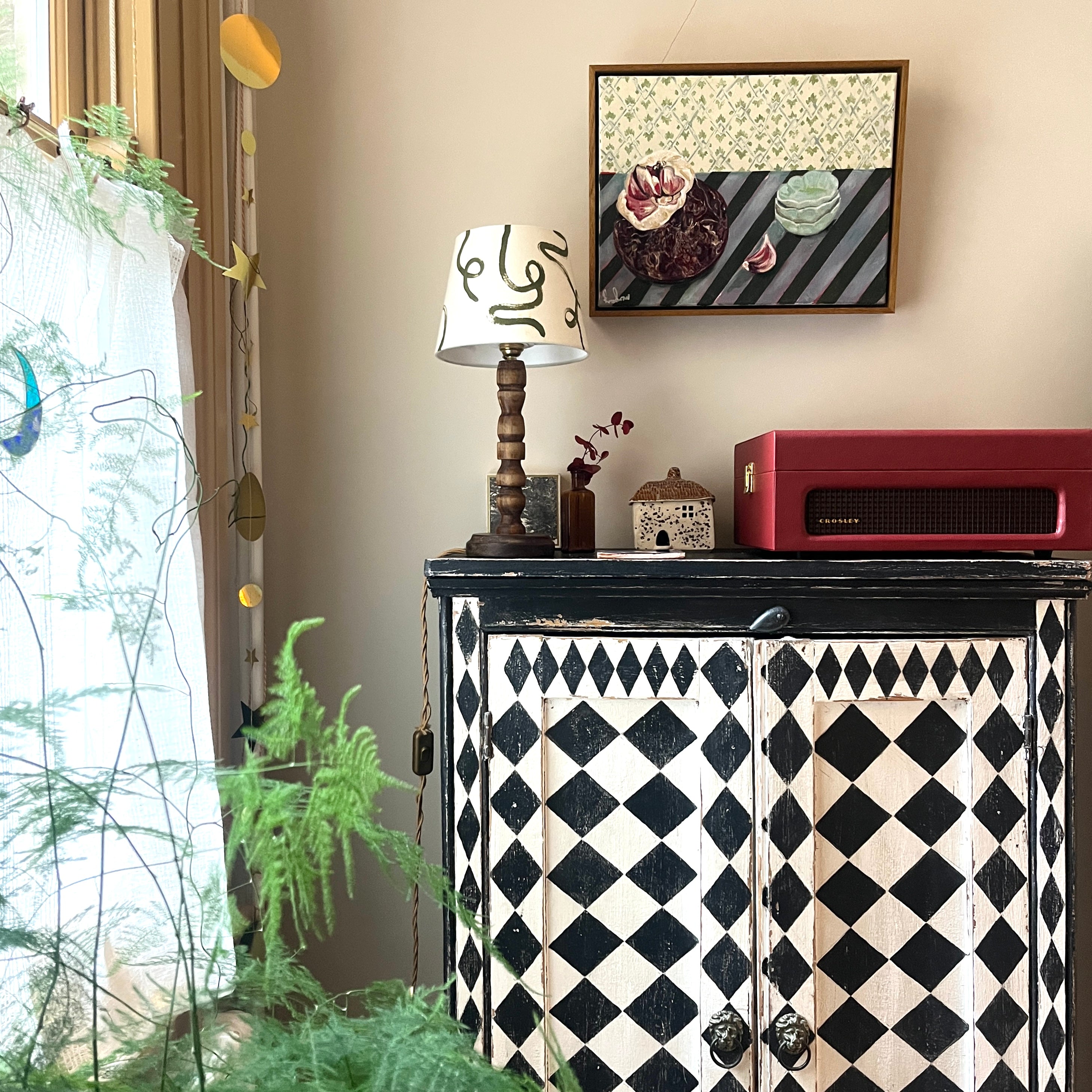 Decorative cabinet with black and white pattern, lamp, and framed picture in a room with plants.