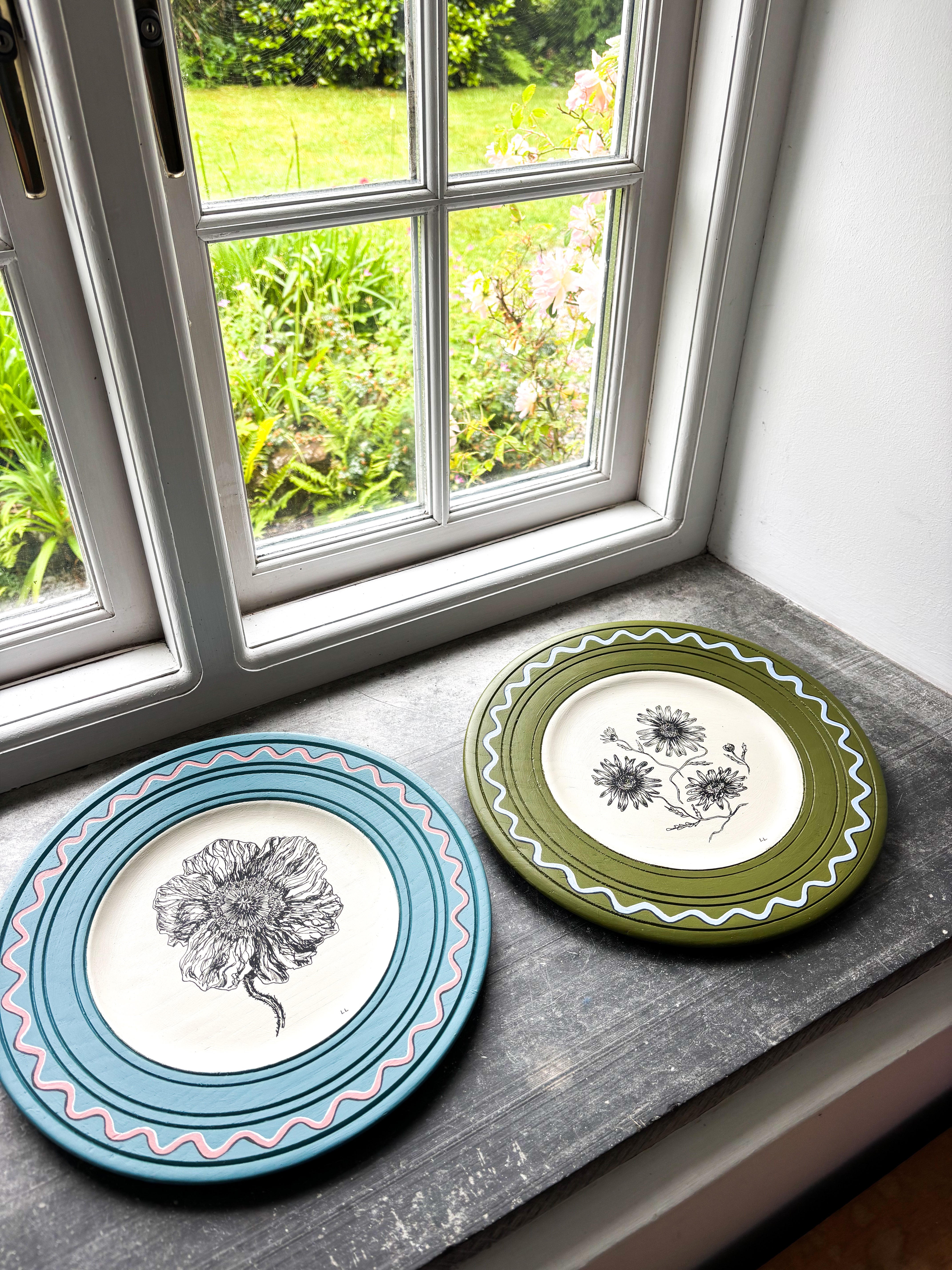 Two decorative plates with floral designs on a windowsill with a view of greenery outside.