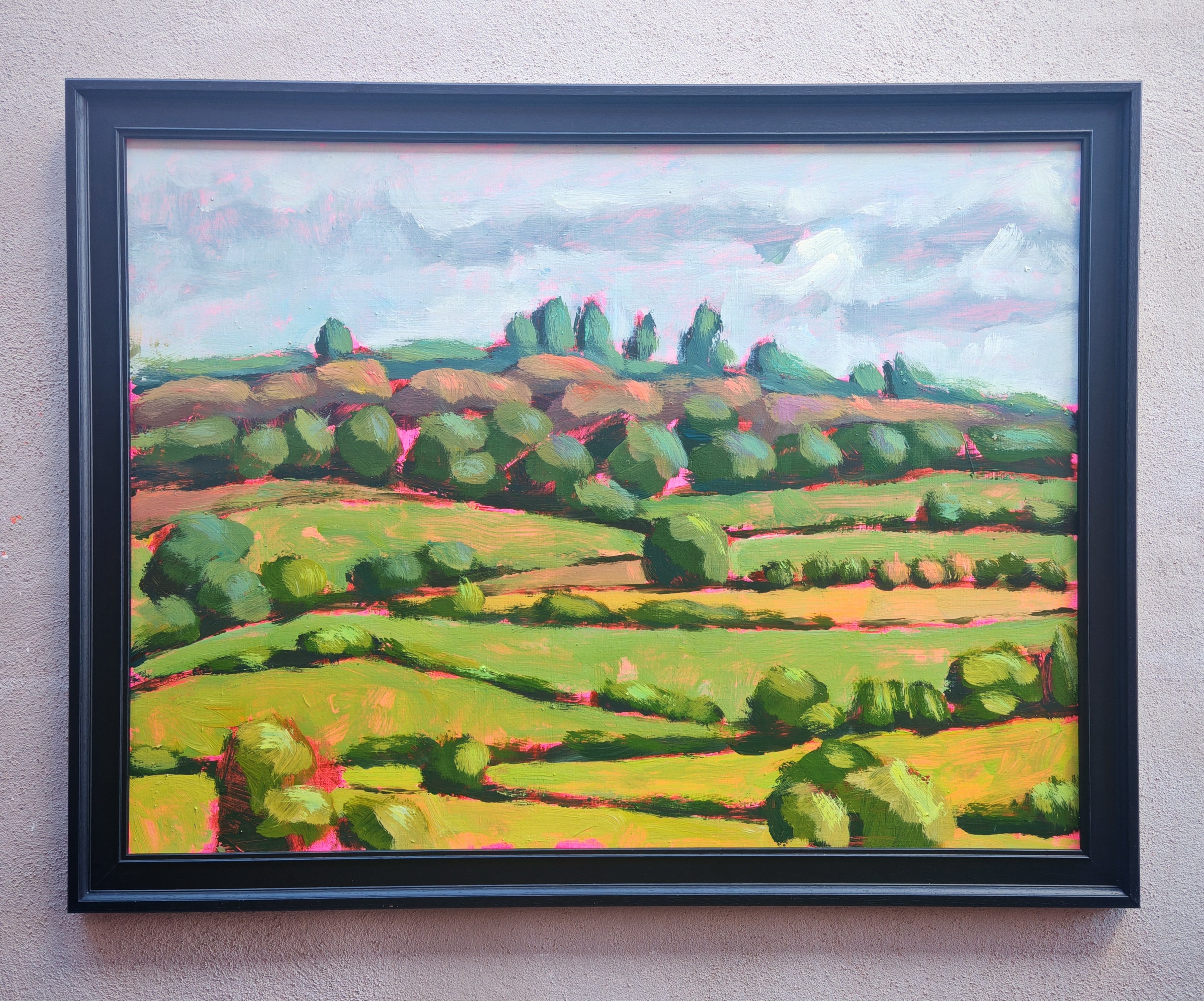 A framed landscape oil painting featuring rolling green and yellow fields, trees, and a cloudy sky, housed in a black frame and mounted on a textured beige wall.