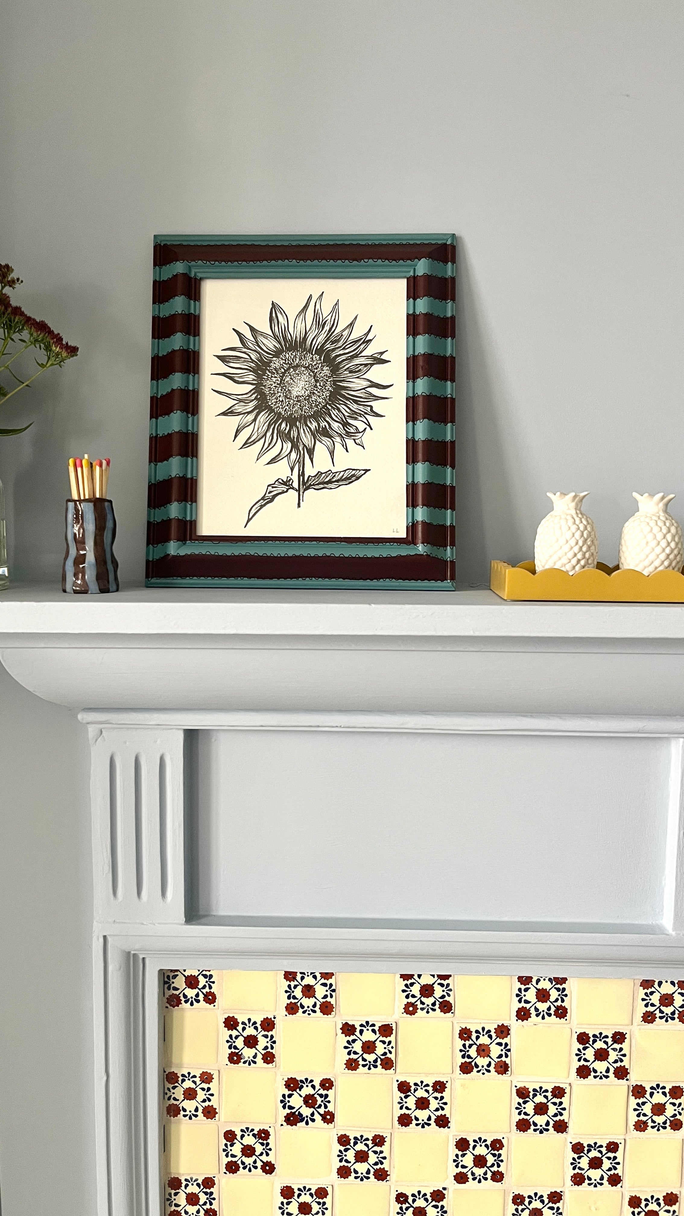 Framed artwork of a sunflower on a white shelf with decorative items.