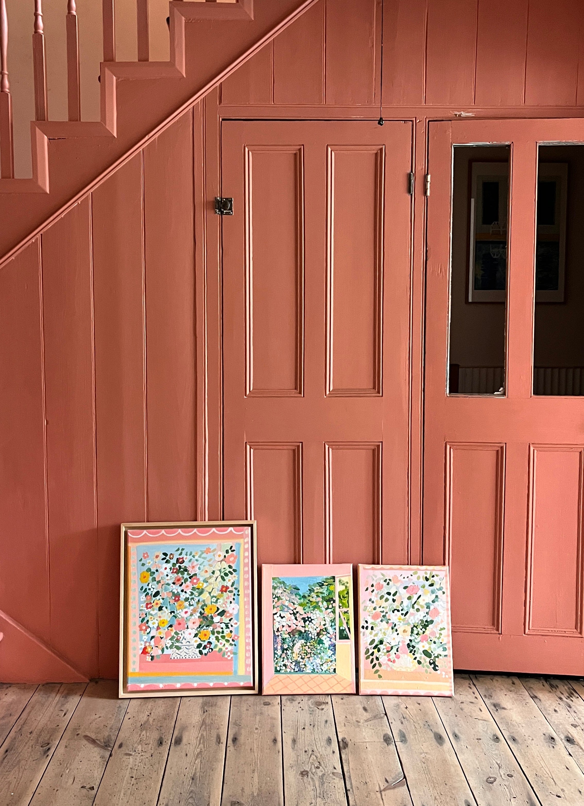 Three framed artworks on a wooden floor with a pink wall and staircase in the background.