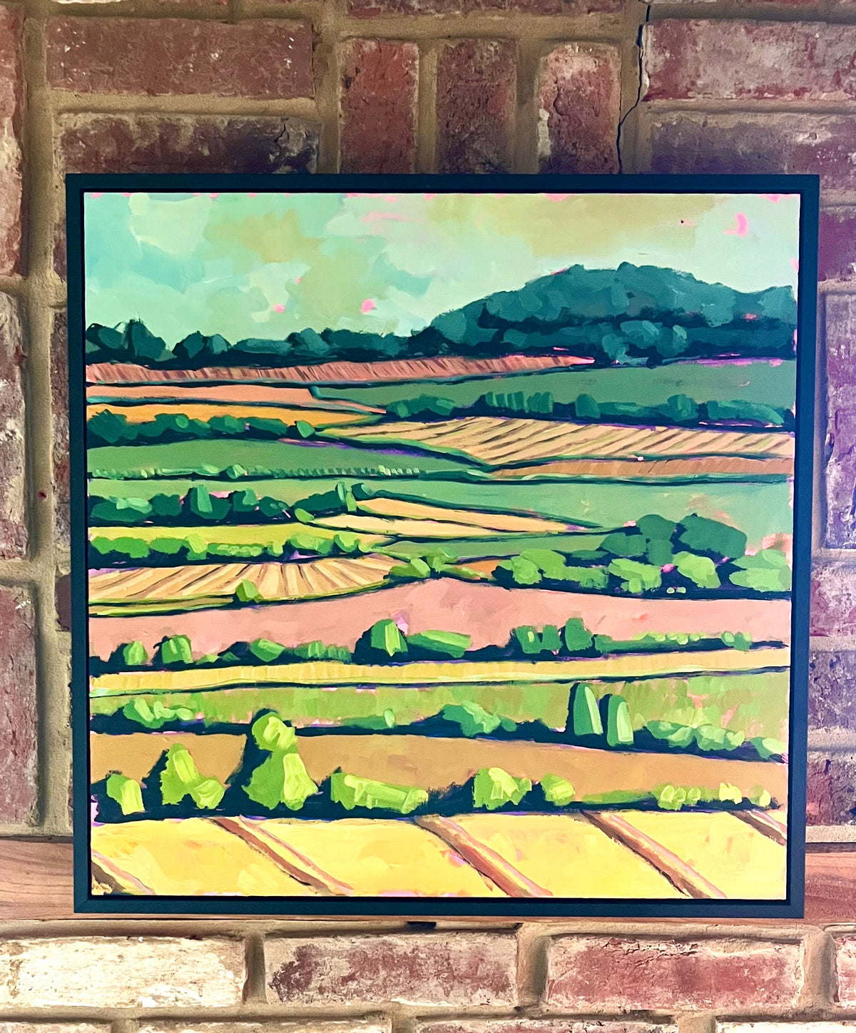 A framed landscape painting of a colorful patchwork field with green hills and a pale sky, mounted on a rustic red and beige brick wall.