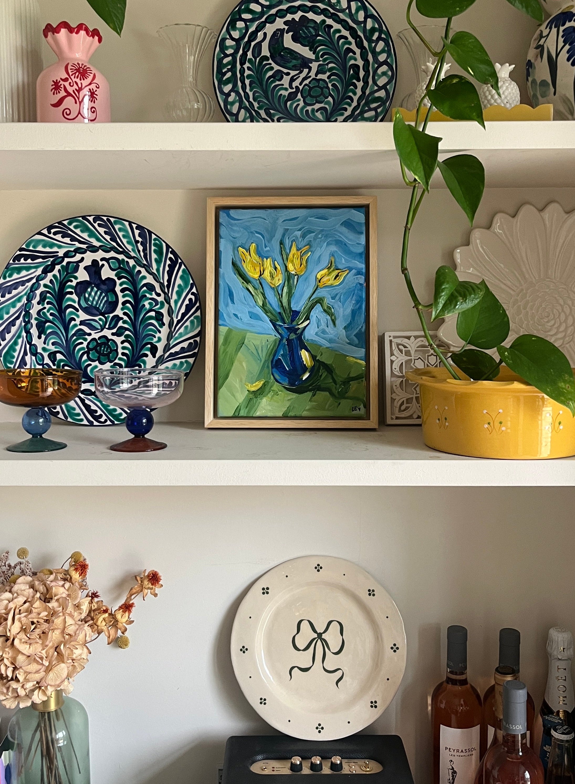 Decorative shelf with plates, a painting, and plants against a white wall.