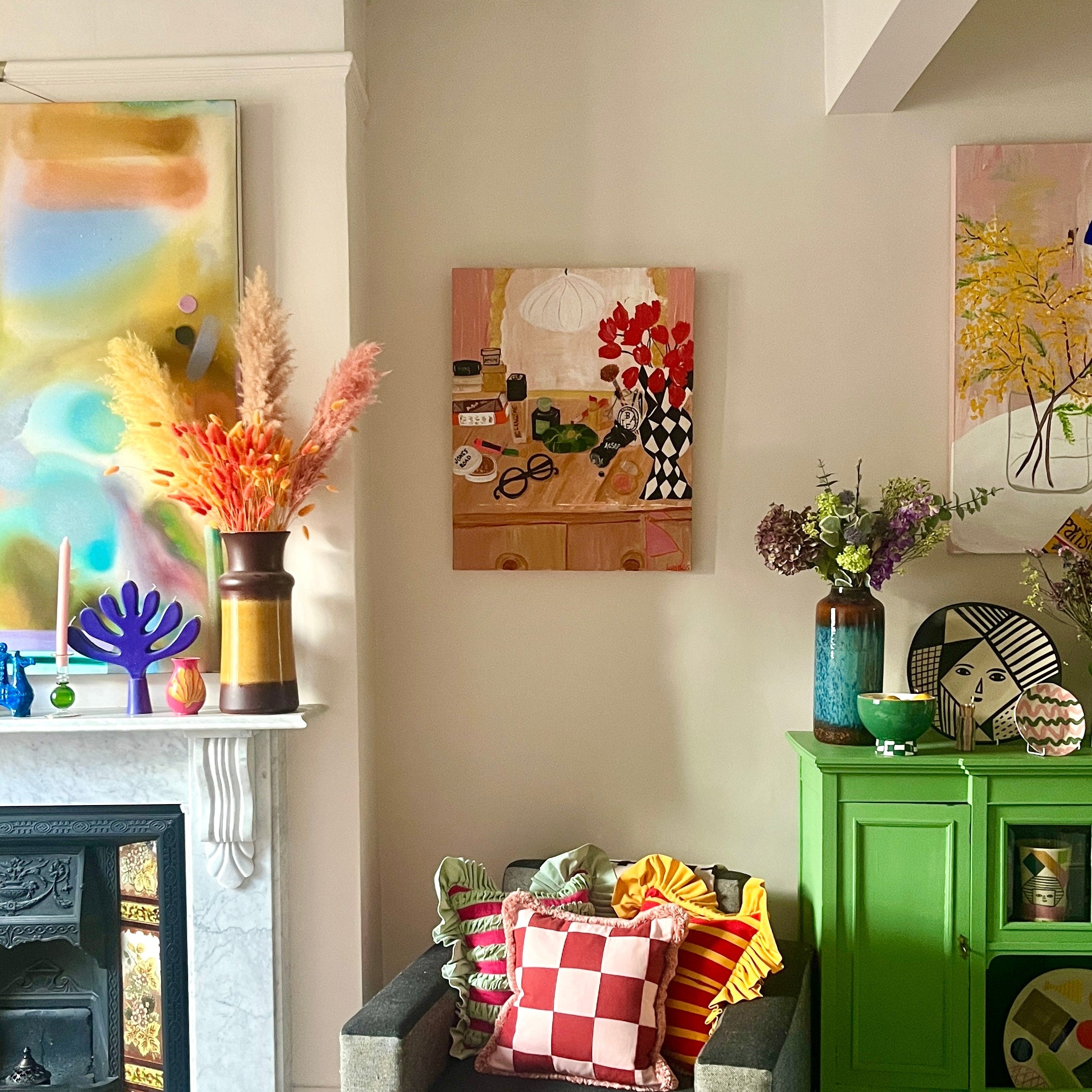 Picture of a dressing table above a chair with bright cushions , green unit and fireplace.