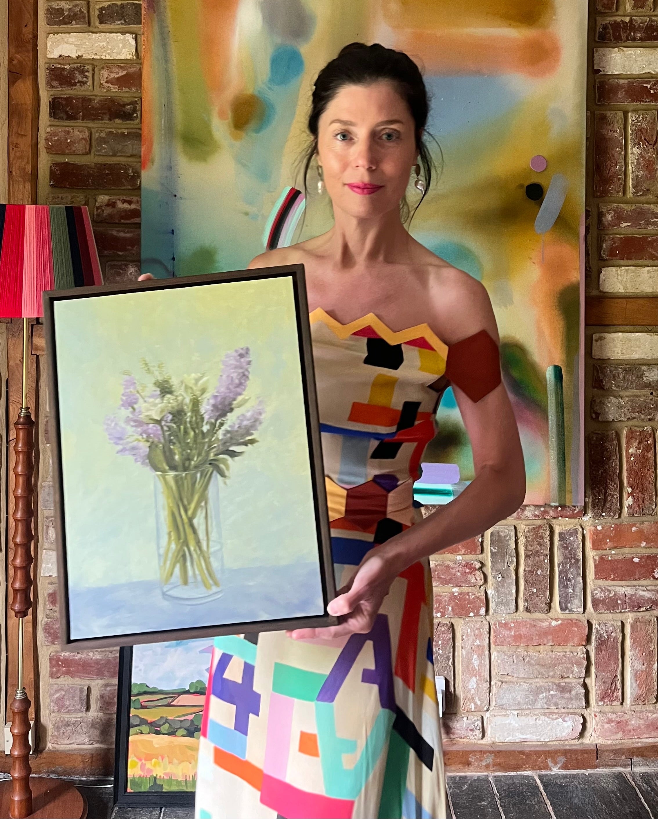 Woman holding a framed painting of flowers in front of a colorful abstract painting on a brick wall.