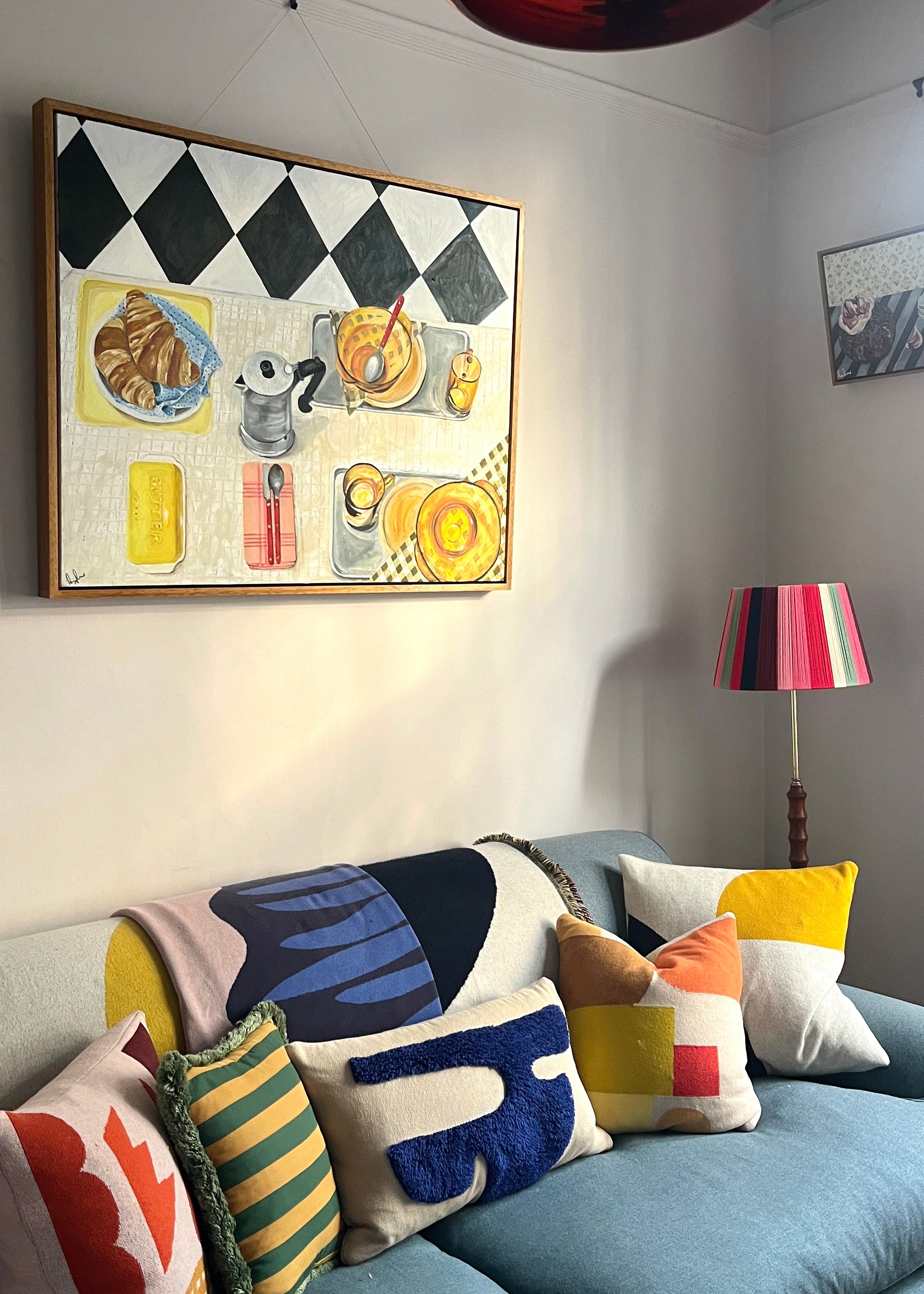 Colorful cushions on a sofa with a decorative Emma Gurner painting of a breakfast scene and lamp in the background.