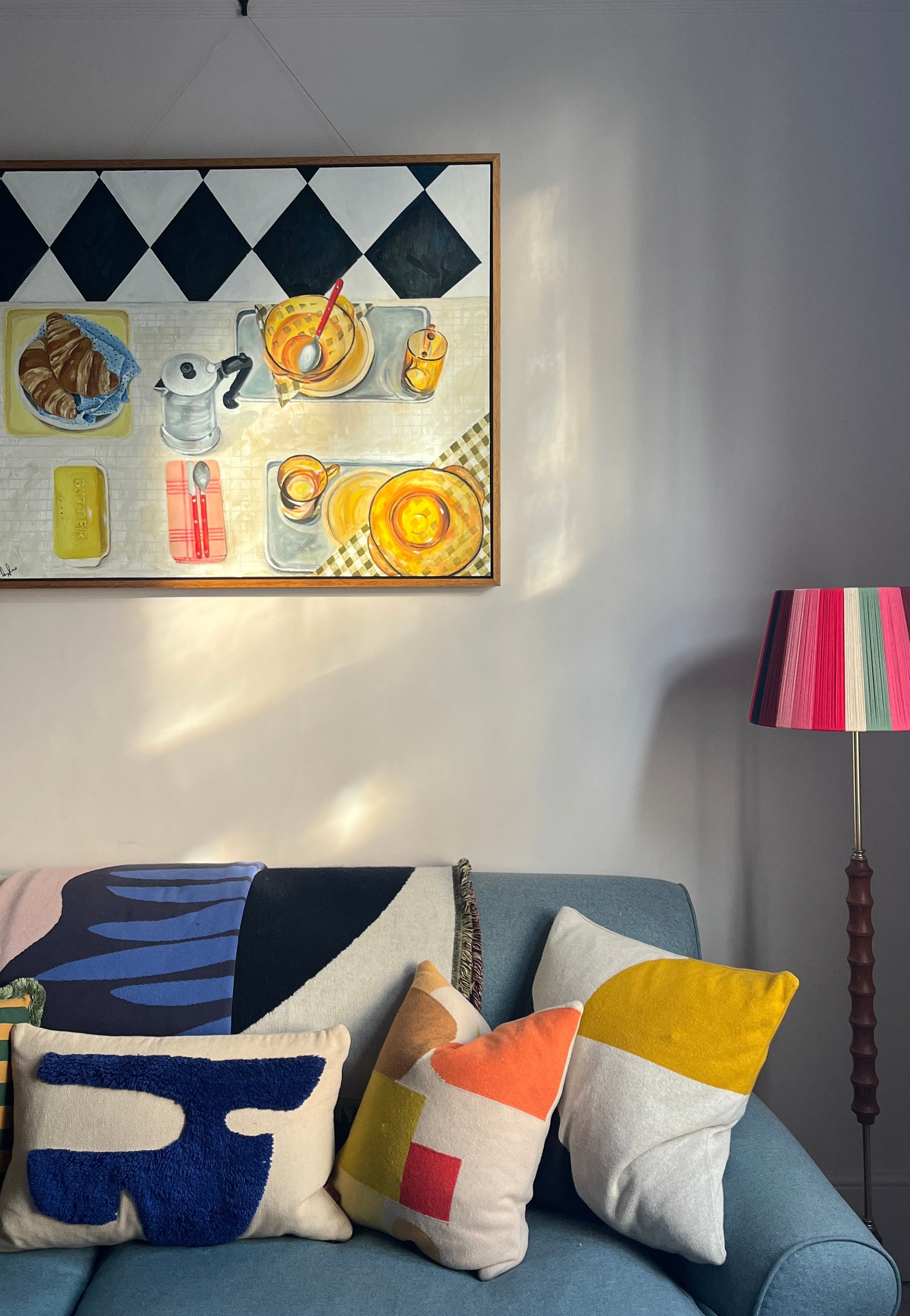 Colorful pillows on a blue sofa with a framed Emma Gurner artwork depicting a breakfast scene and lamp in the background.