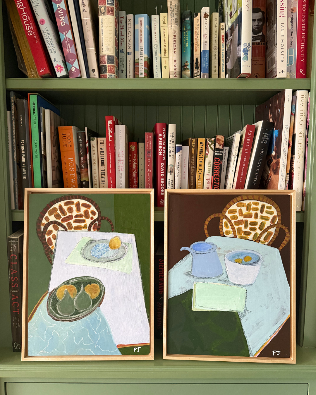 Two framed paintings of table settings on a bookshelf with books.
