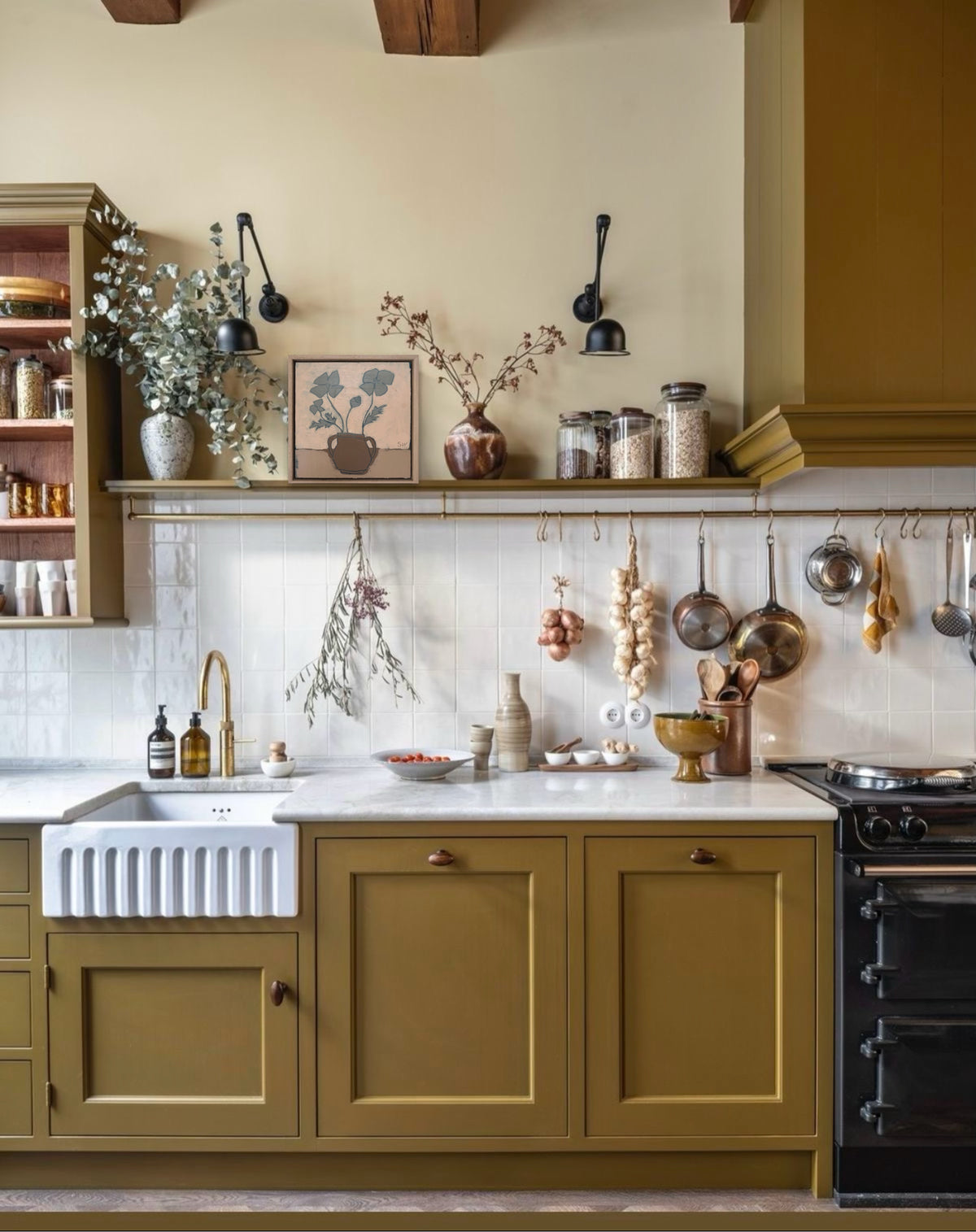 A styled kitchen interior with olive green cabinets, a white farmhouse sink, white marble countertop, hanging copper pots and utensils, and decorative dried flowers and jars on open shelves.