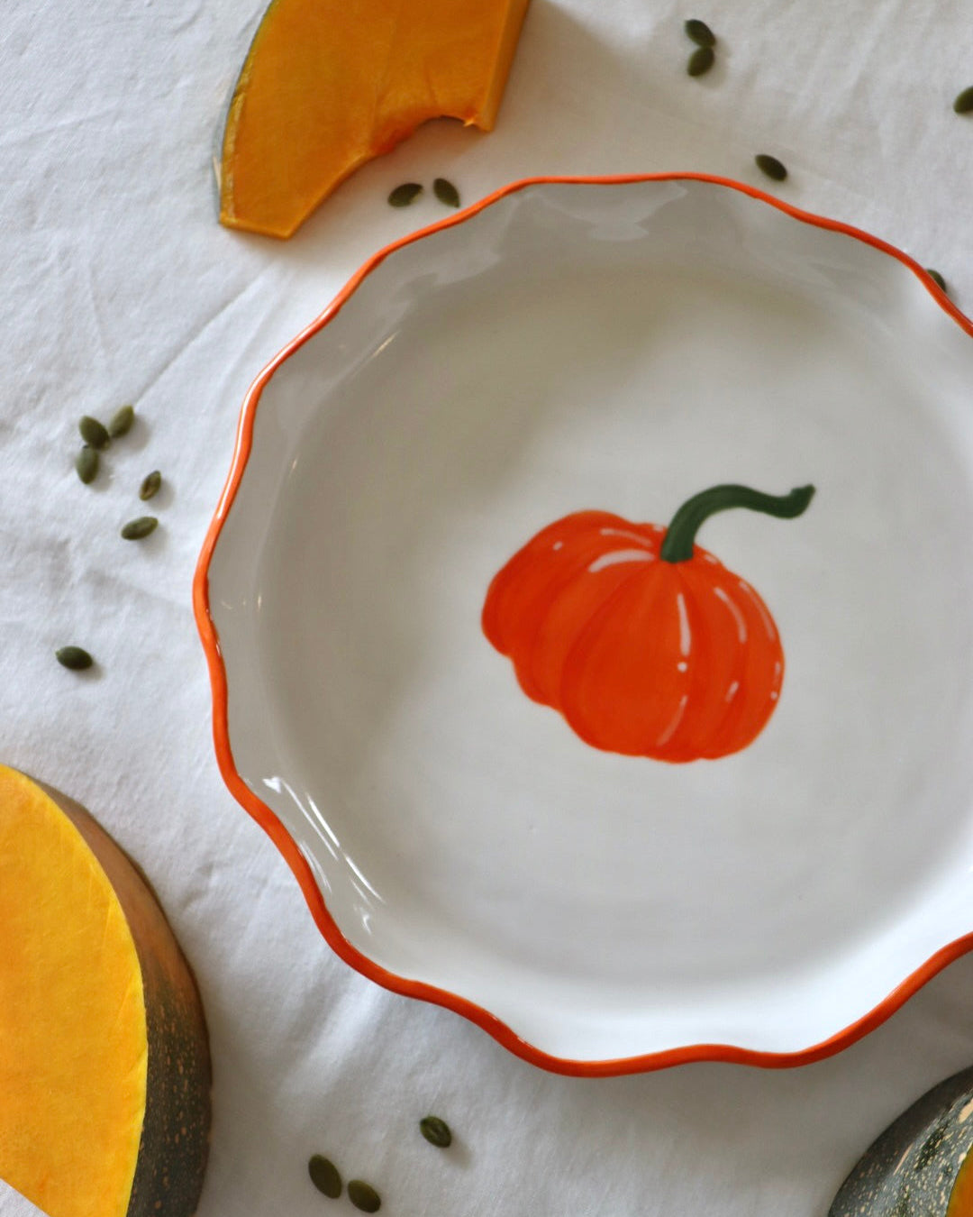 A white ceramic bowl with a scalloped rim, featuring a painted orange pumpkin in the center and an orange border. The bowl is styled on a white fabric background with pumpkin slices and seeds.