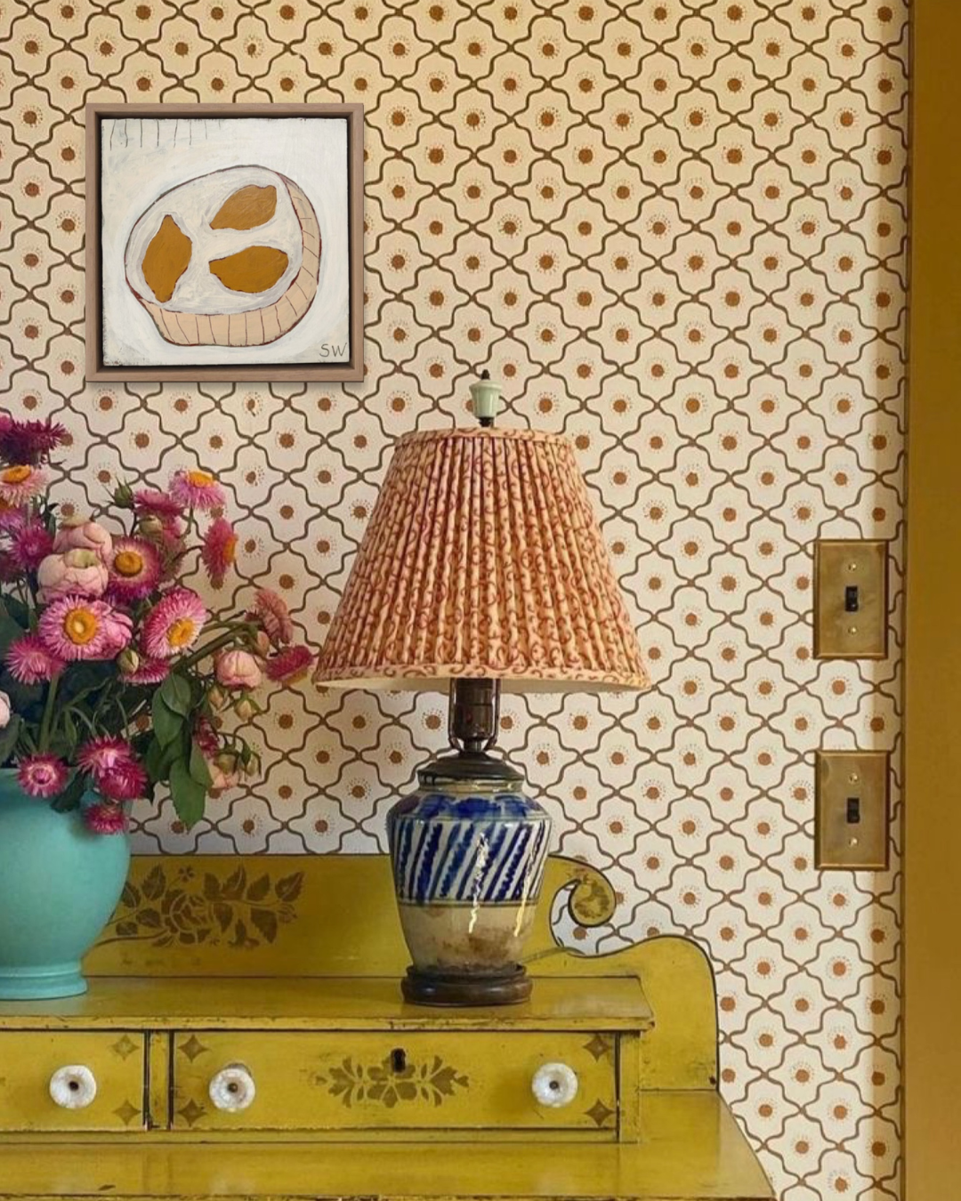 A vintage-style table lamp with a textured peach-pink pleated shade and a blue-and-white striped ceramic base sits on a yellow-green dresser against a patterned wallpaper background. A turquoise flower vase with pink flowers is on the left, and a framed art print hangs on the wall.
