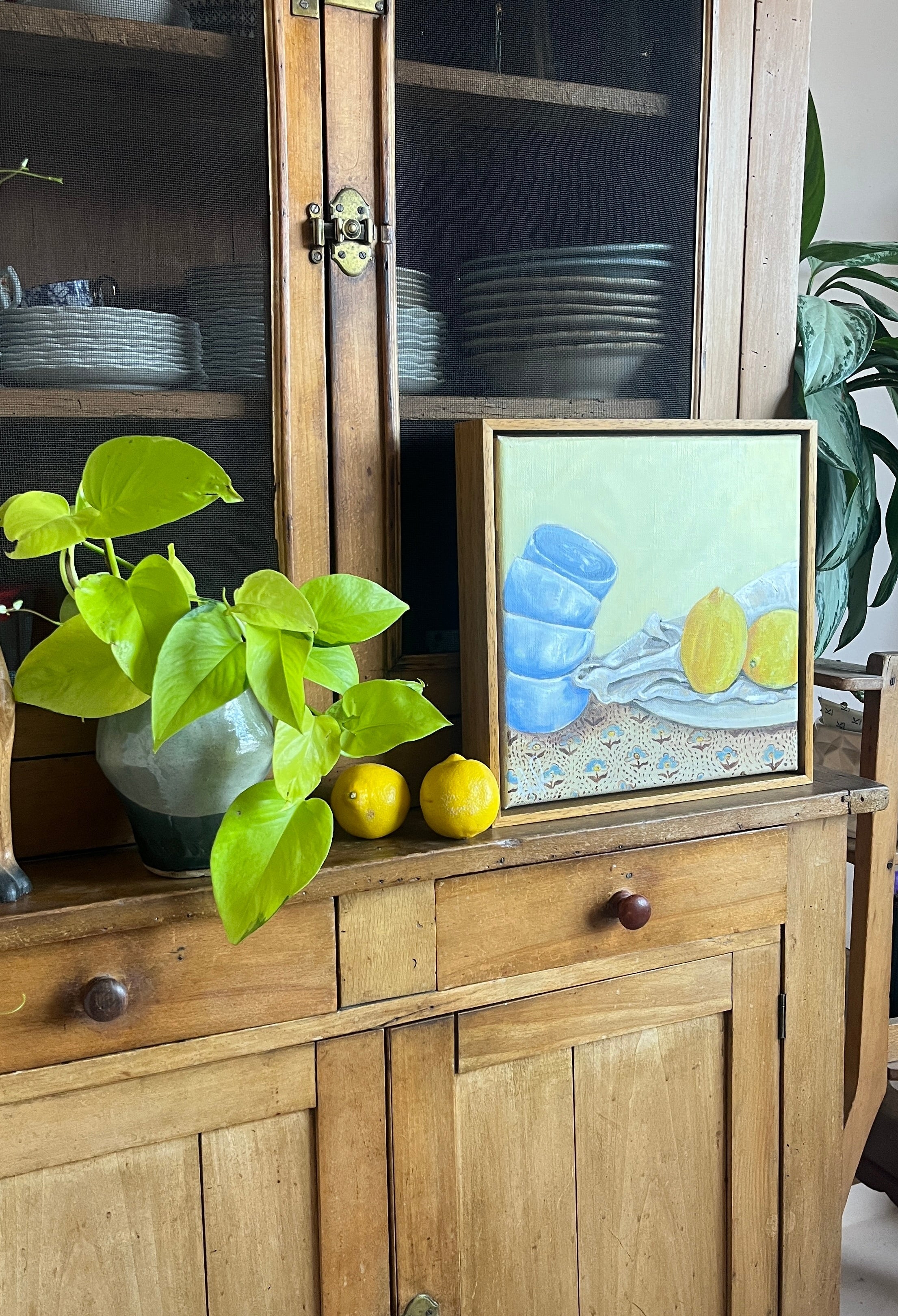 Wooden cabinet with a painting of Emma Gurner lemons and a plant on top