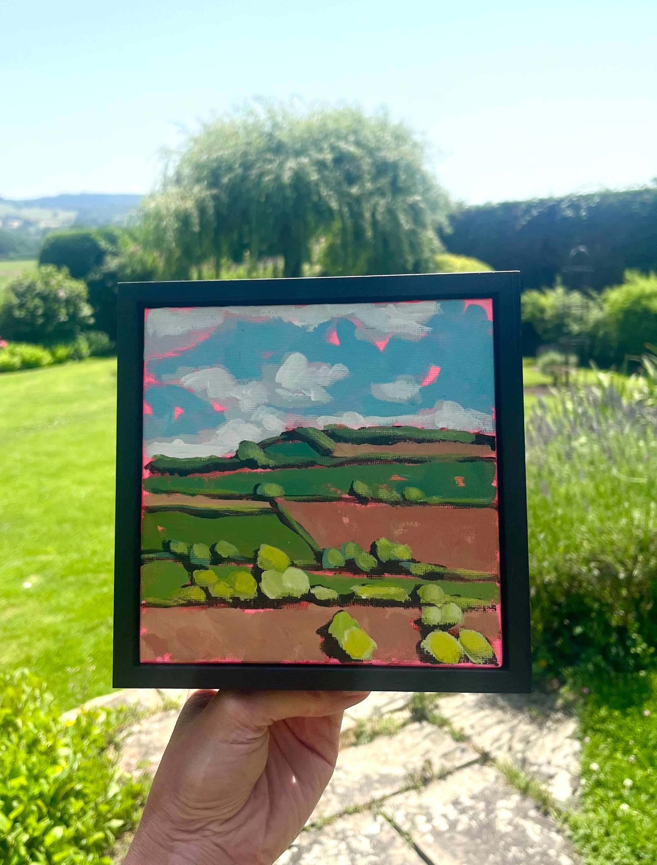Hand holding a small framed painting of a landscape with green fields and blue sky.