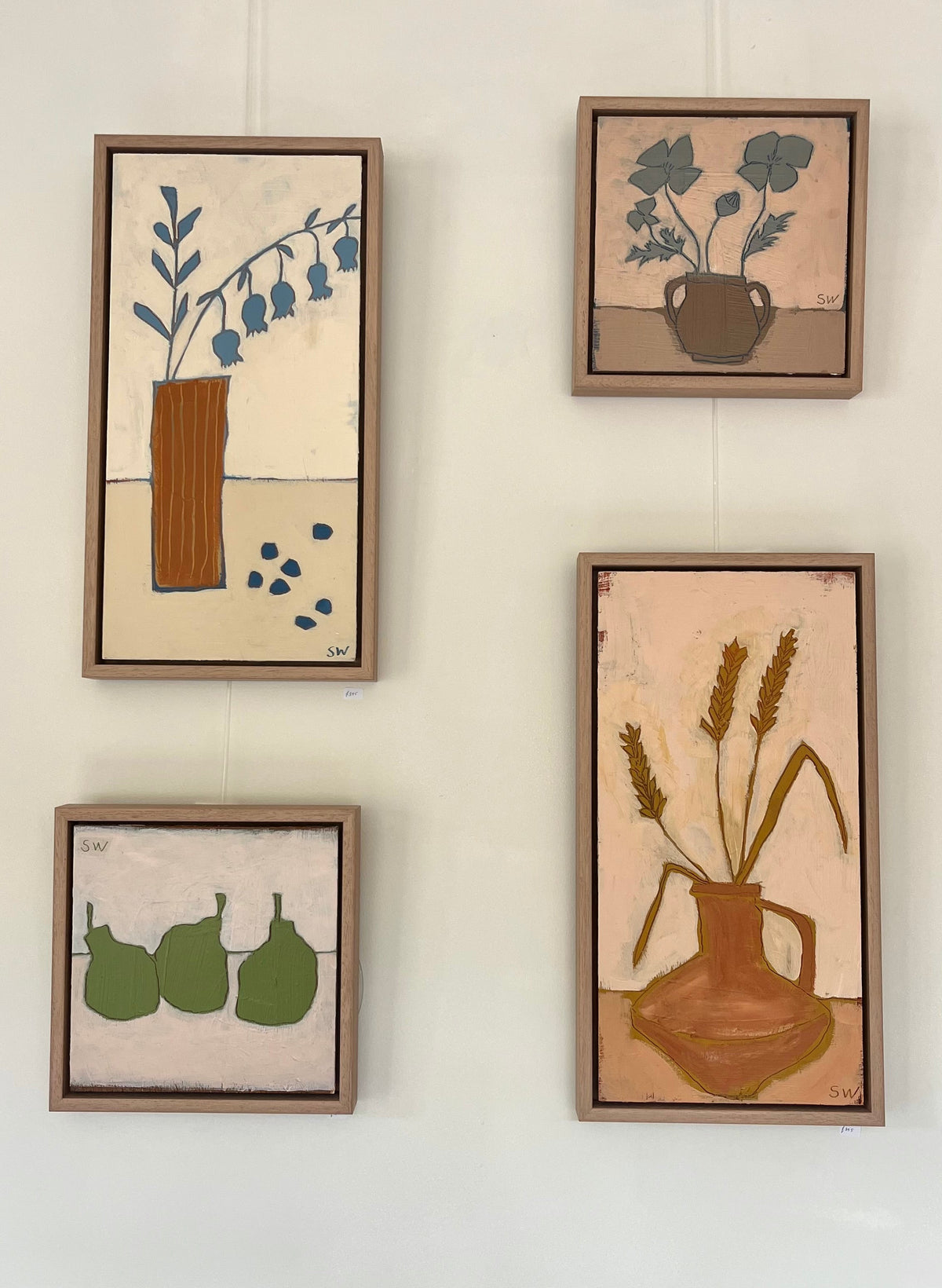 Four framed still life paintings with muted earth tones and blue accents hang on a white wall.