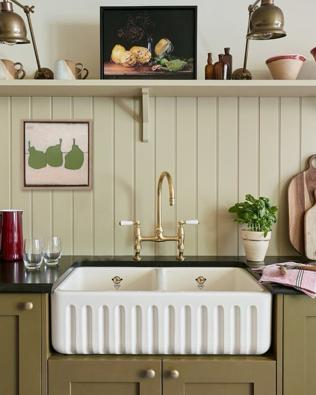 A white farmhouse double-bowl sink with a ribbed front and a polished brass faucet, installed in a kitchen with olive green cabinets, a dark countertop, and decorative wall art.