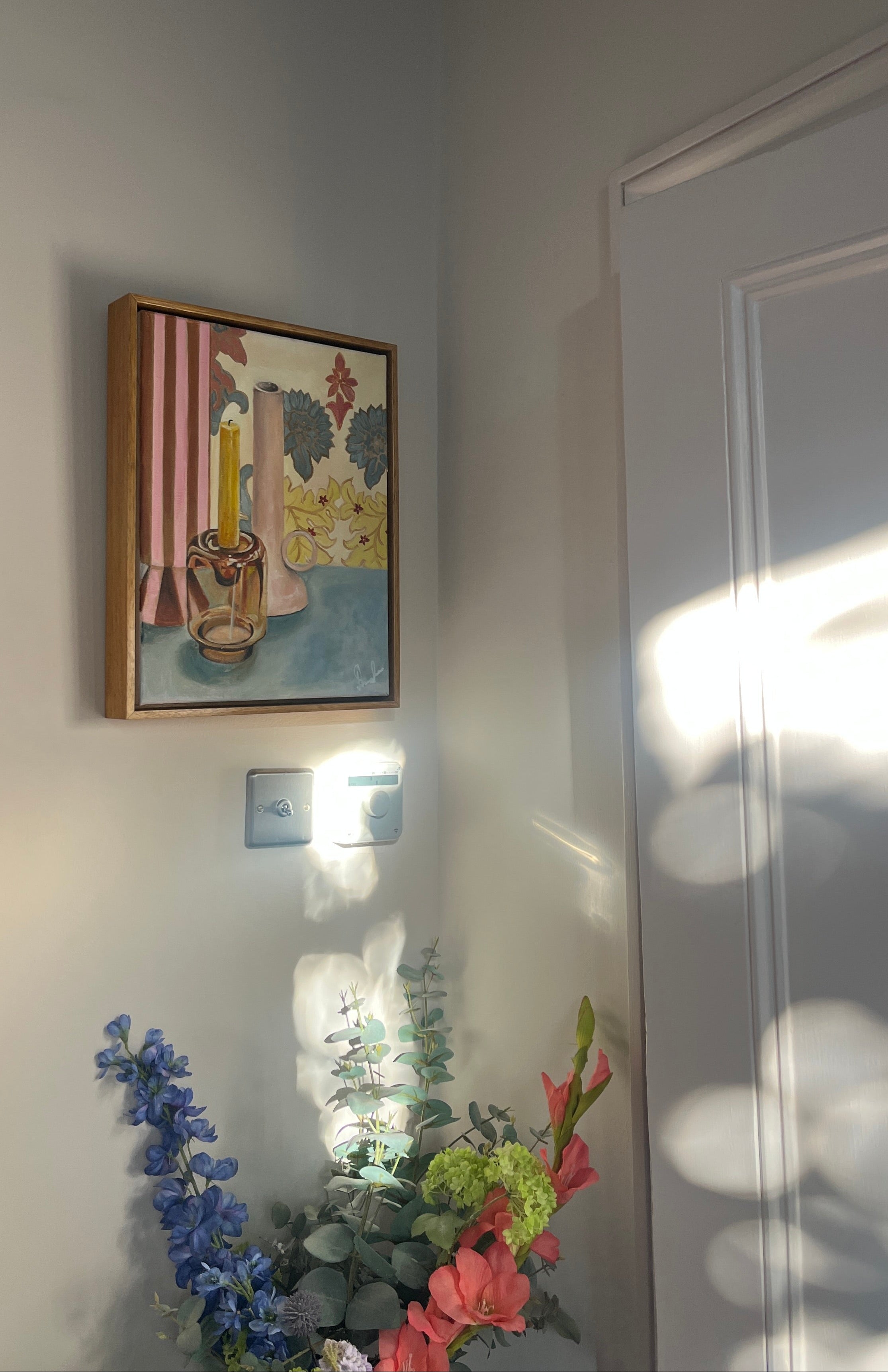 Framed Emma Gurner artwork on a wall with a light fixture and flowers in the foreground.