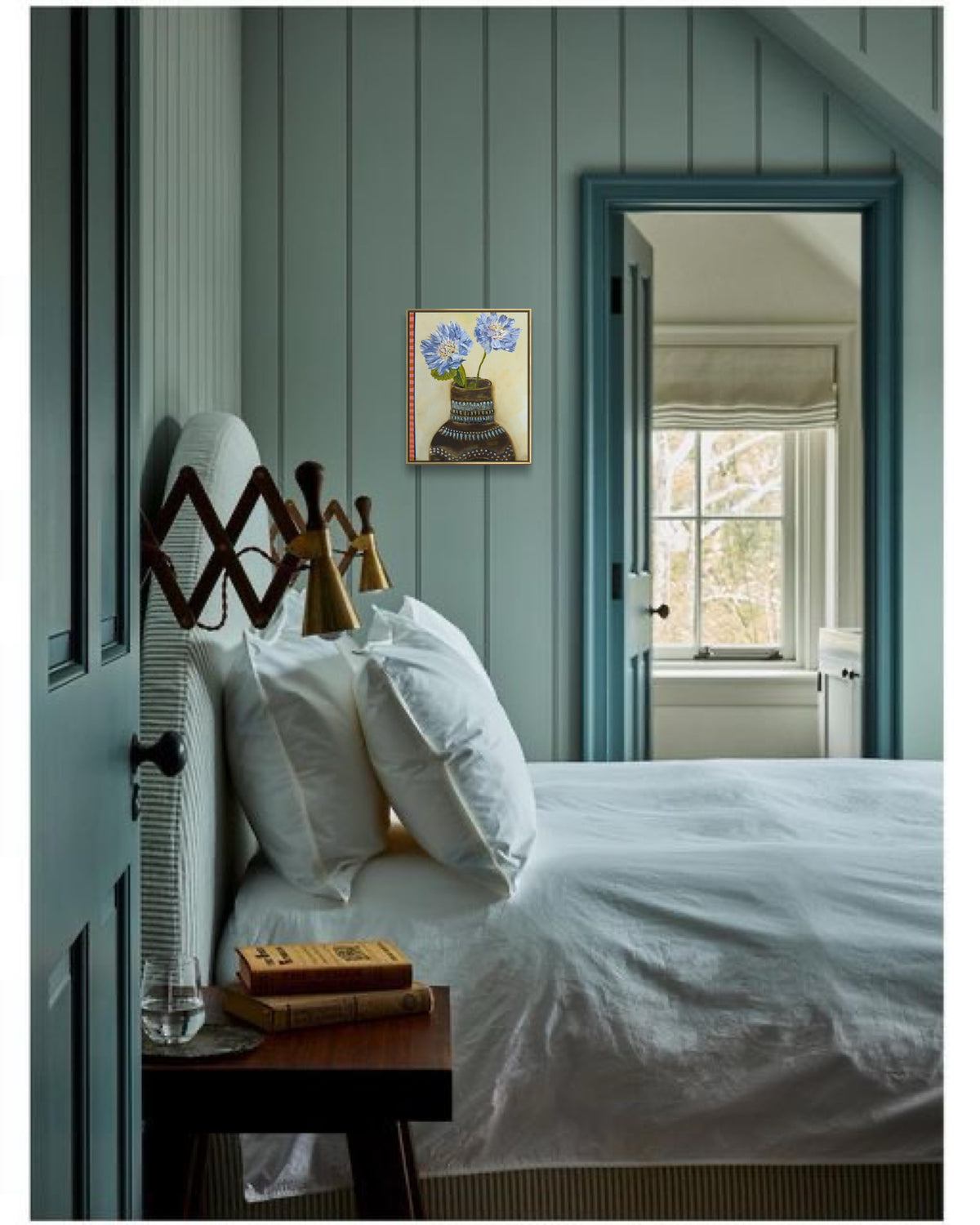 A styled bedroom interior with a bed dressed in white linens, blue paneled walls, a blue door frame, and a small framed floral painting on the wall.