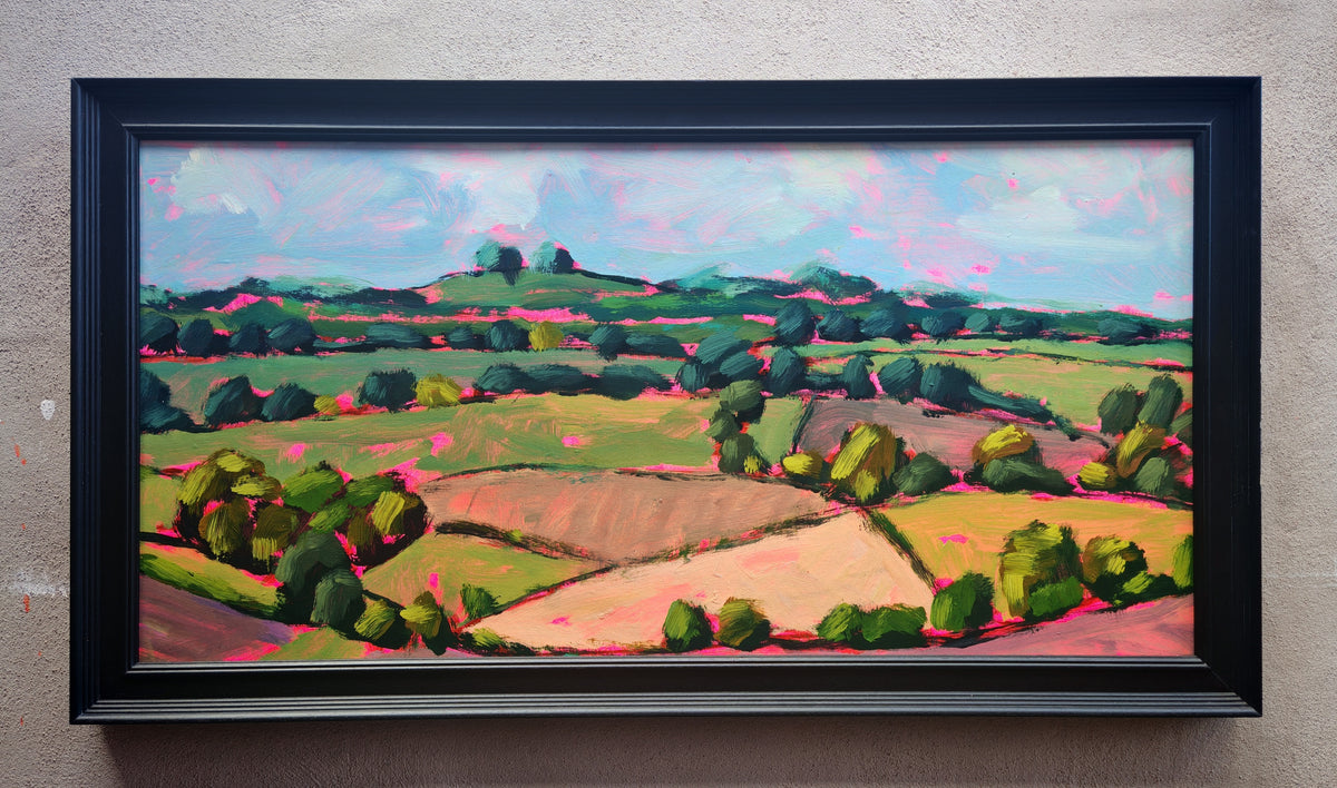 A framed landscape oil painting featuring vibrant green fields, rolling hills, and a blue sky with pinkish clouds, housed in a thick black frame mounted on a textured beige wall.