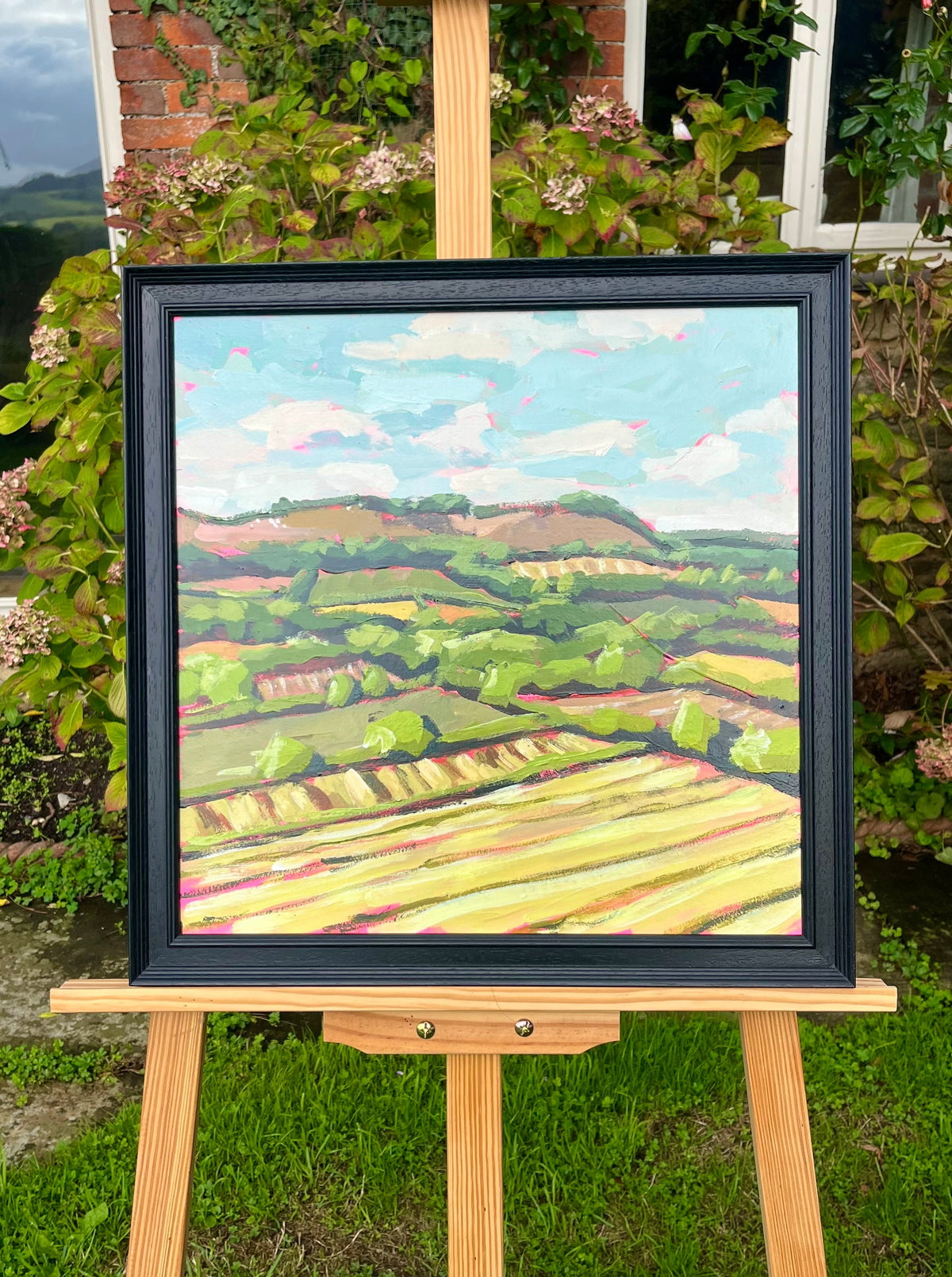 A framed landscape oil painting on an easel in a garden setting. The painting depicts rolling green hills, yellow fields, and a blue sky, housed in a black frame.