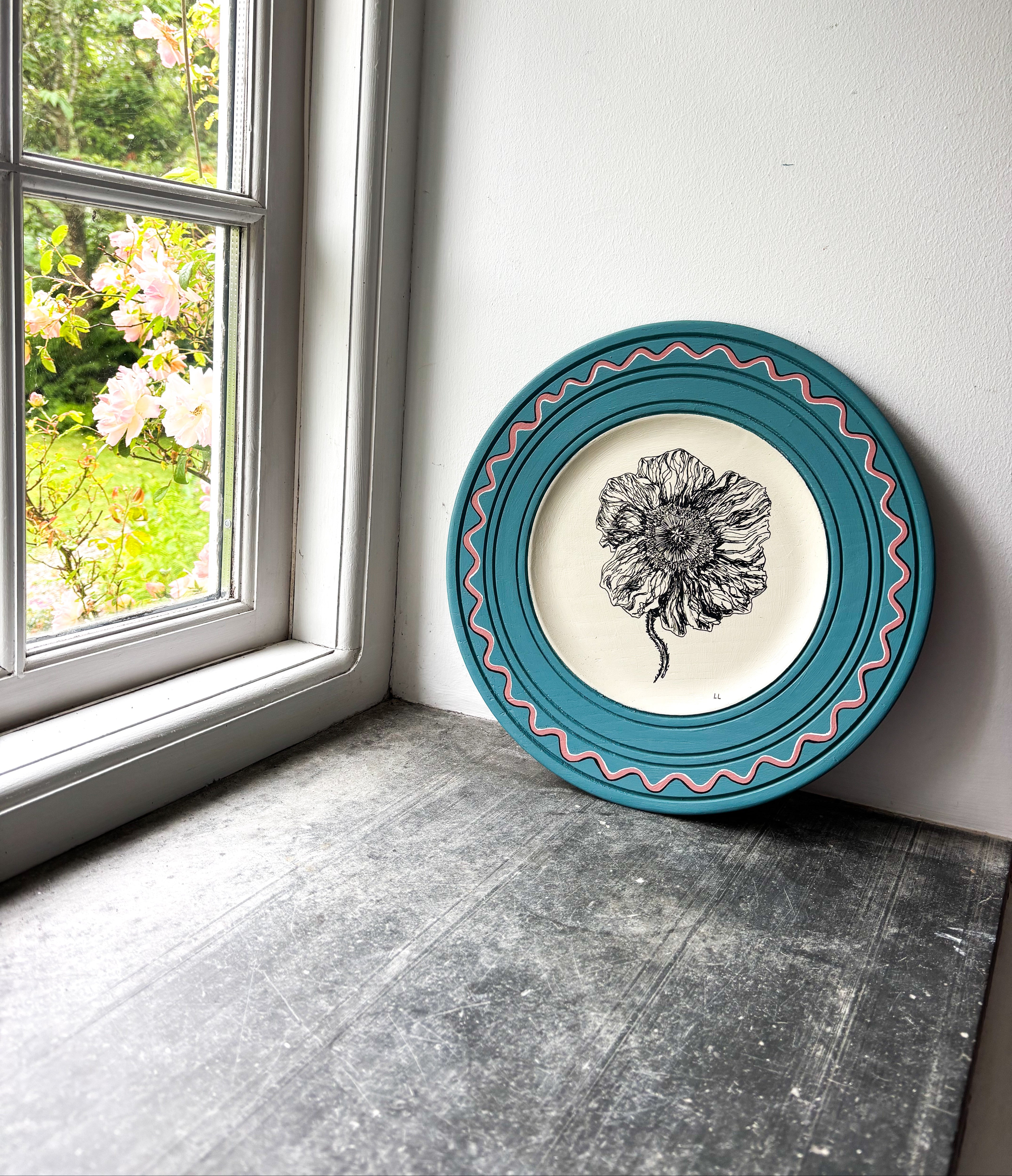 Decorative plate with floral design on a wall next to a window with greenery outside