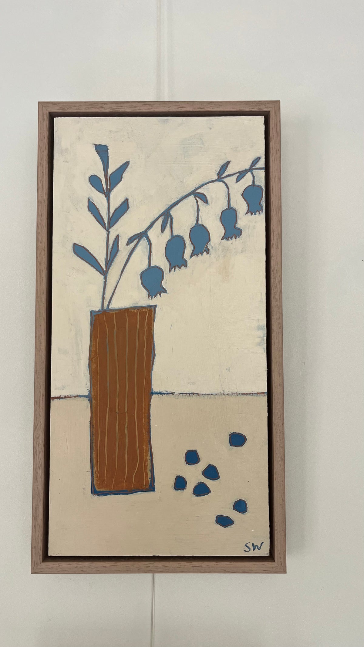 A framed vertical painting featuring a brown vase with blue bell-shaped flowers on a cream background, hung on a white wall.