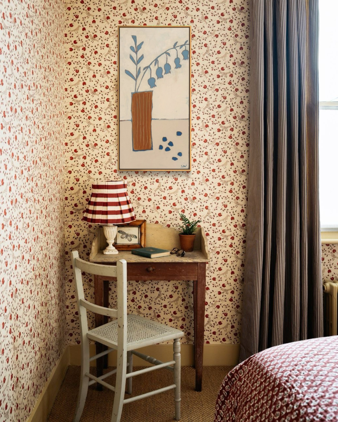 A cozy bedroom corner featuring floral wallpaper with a red and cream pattern, a small wooden desk with a red-and-white striped lamp, a white chair, and a framed botanical print on the wall.