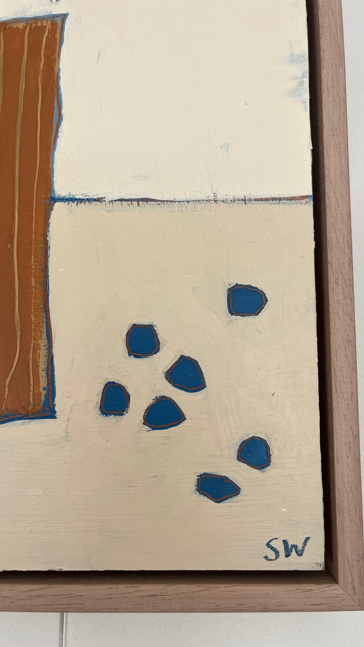 Close-up of an abstract framed painting with cream and beige sections, a vertical brown block on the left, and several blue organic shapes on the lower right. The artist signature 'SW' is visible in the bottom corner.