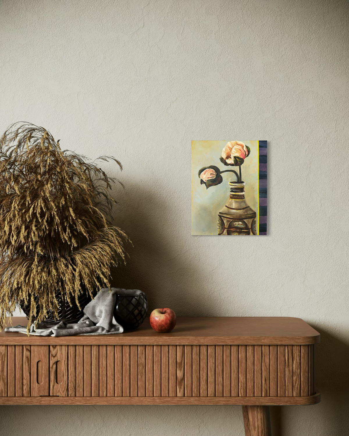 A small framed art print depicting a surreal flower arrangement in a vase, mounted on a textured beige wall above a wooden console table with a dried plant, a black bag, and a red apple.