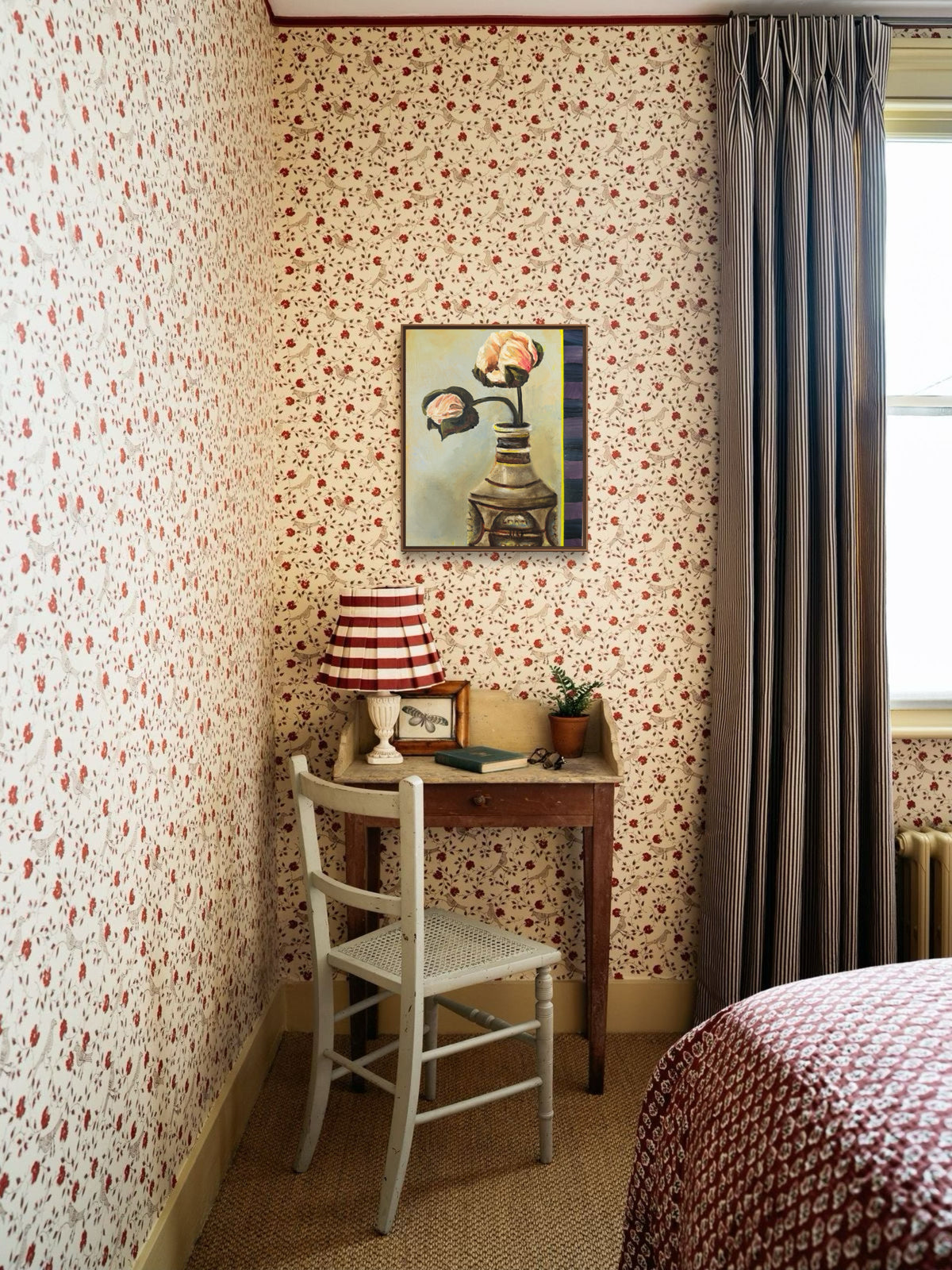 A vintage-style bedroom corner with floral wallpaper, a small wooden desk, a white chair, a red-and-white striped lamp, and a framed painting on the wall. A window with dark gray curtains is visible on the right, along with part of a patterned bedspread.