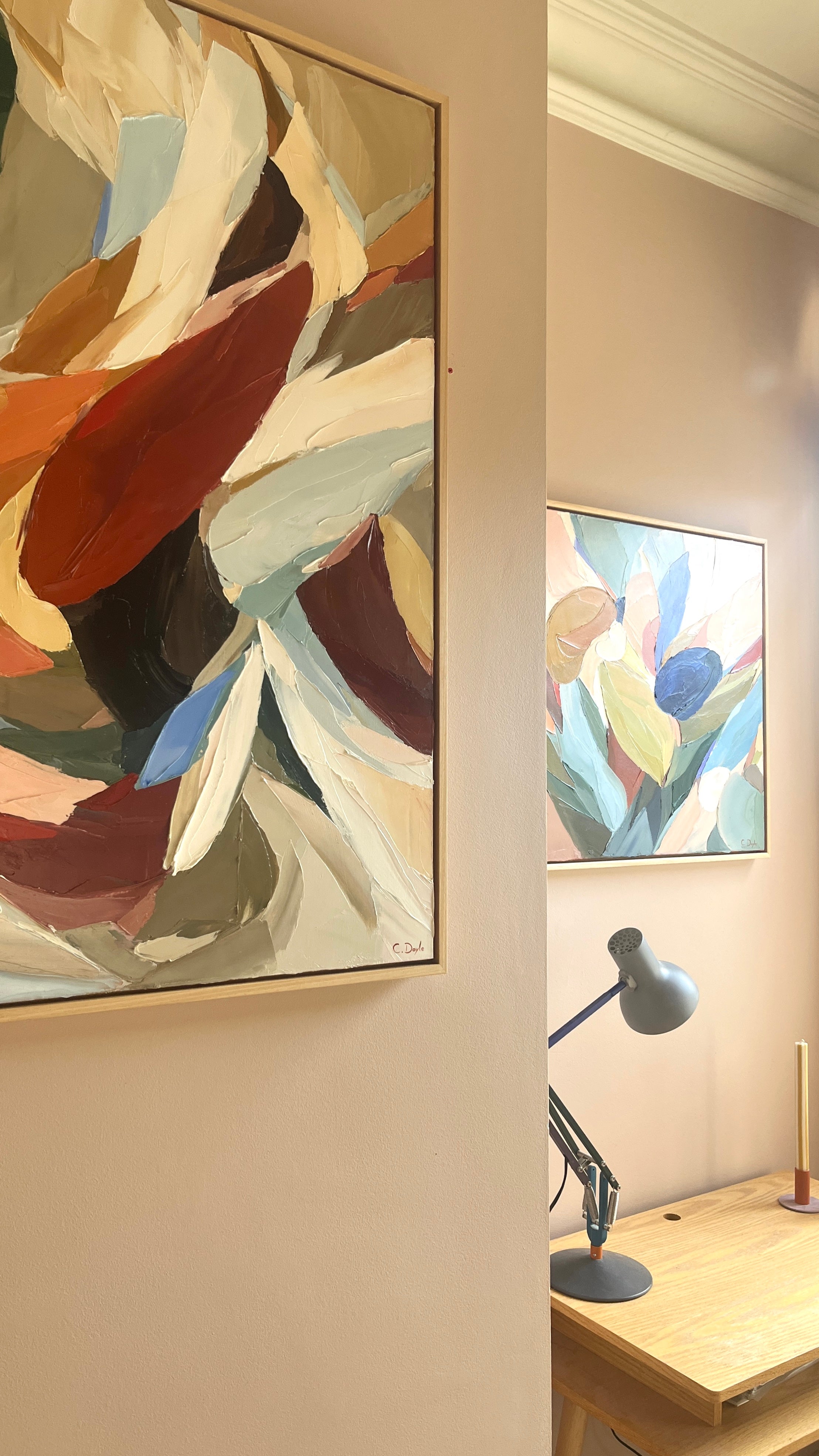 Two abstract artworks on a wall with a desk and lamp in the foreground.