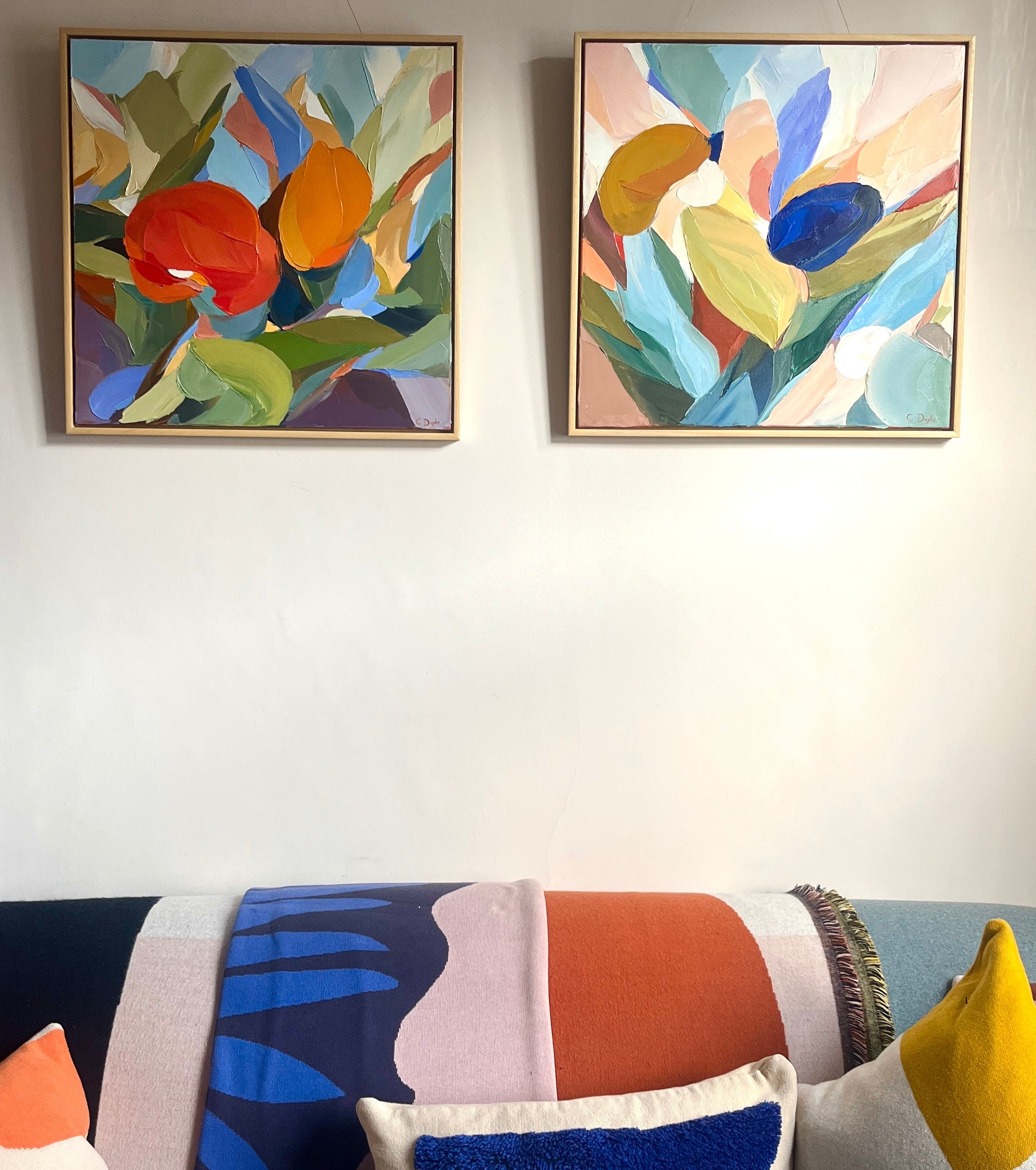 Two abstract paintings on a wall above a colorful sofa with patterned cushions.