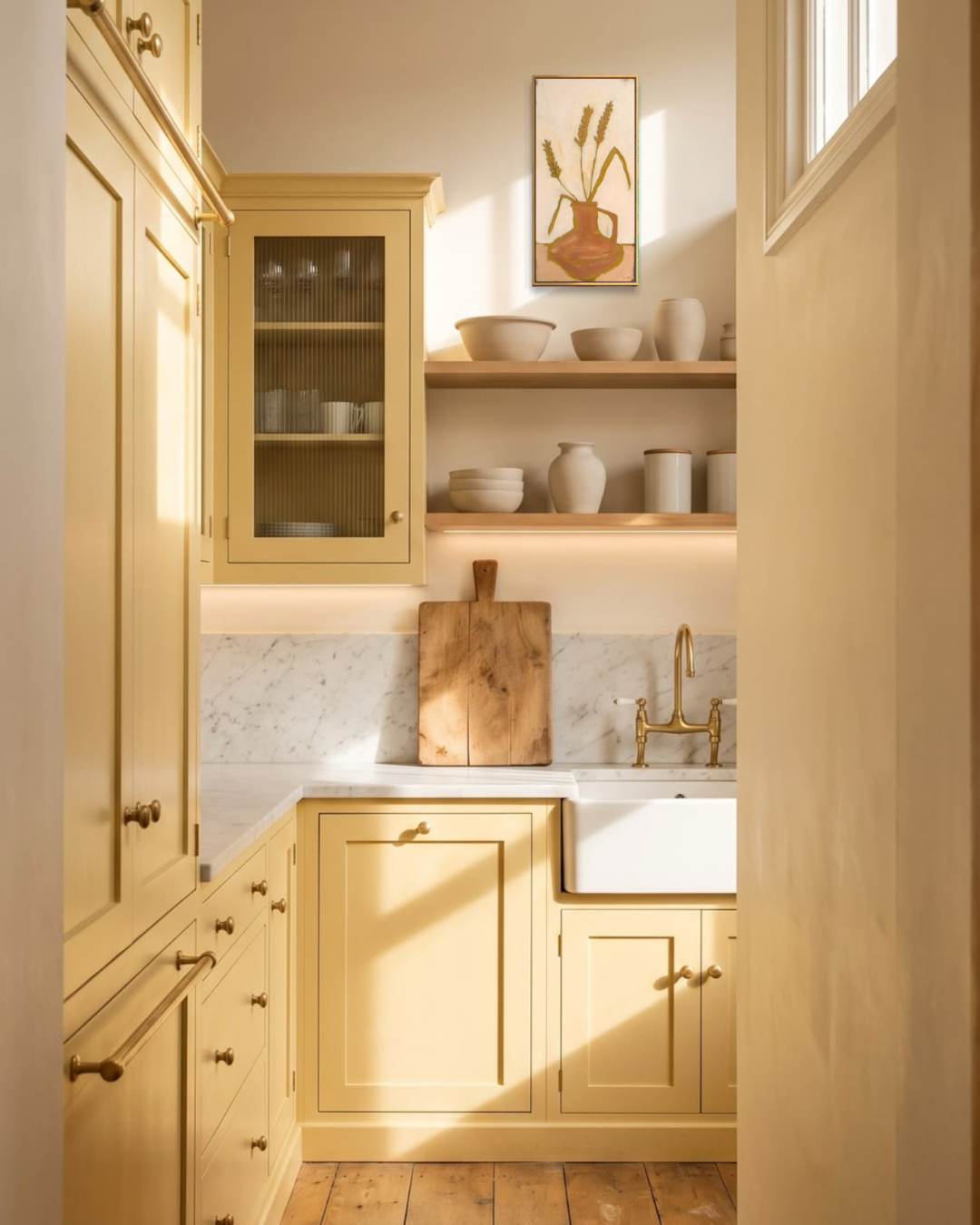 A warm, sunlit kitchen interior with pale yellow cabinets, open wood shelves holding ceramic vases and bowls, a white farmhouse sink, a brass faucet, and a wooden cutting board leaning against the backsplash.
