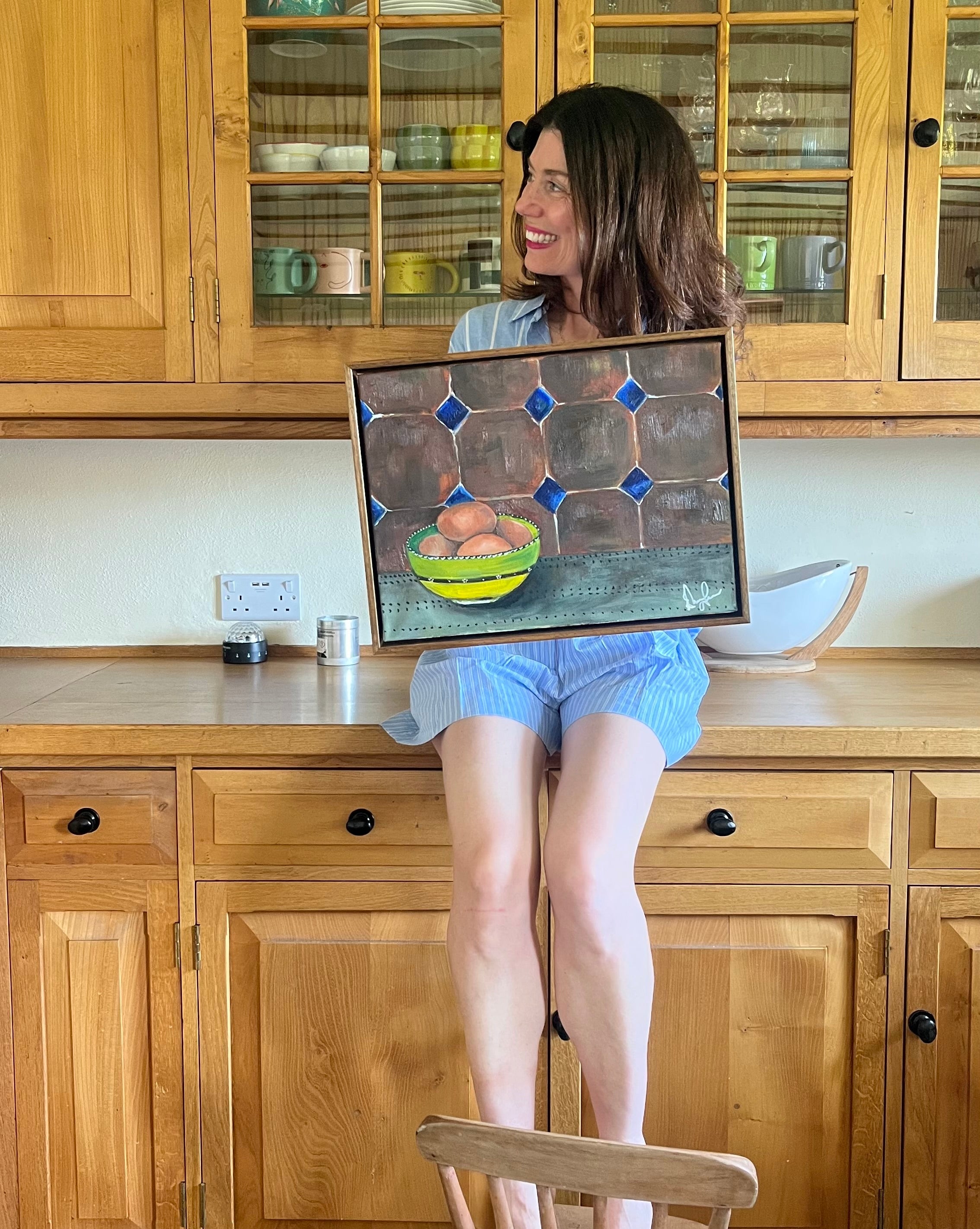 Person holding a framed painting of a bowl with fruit in a kitchen.