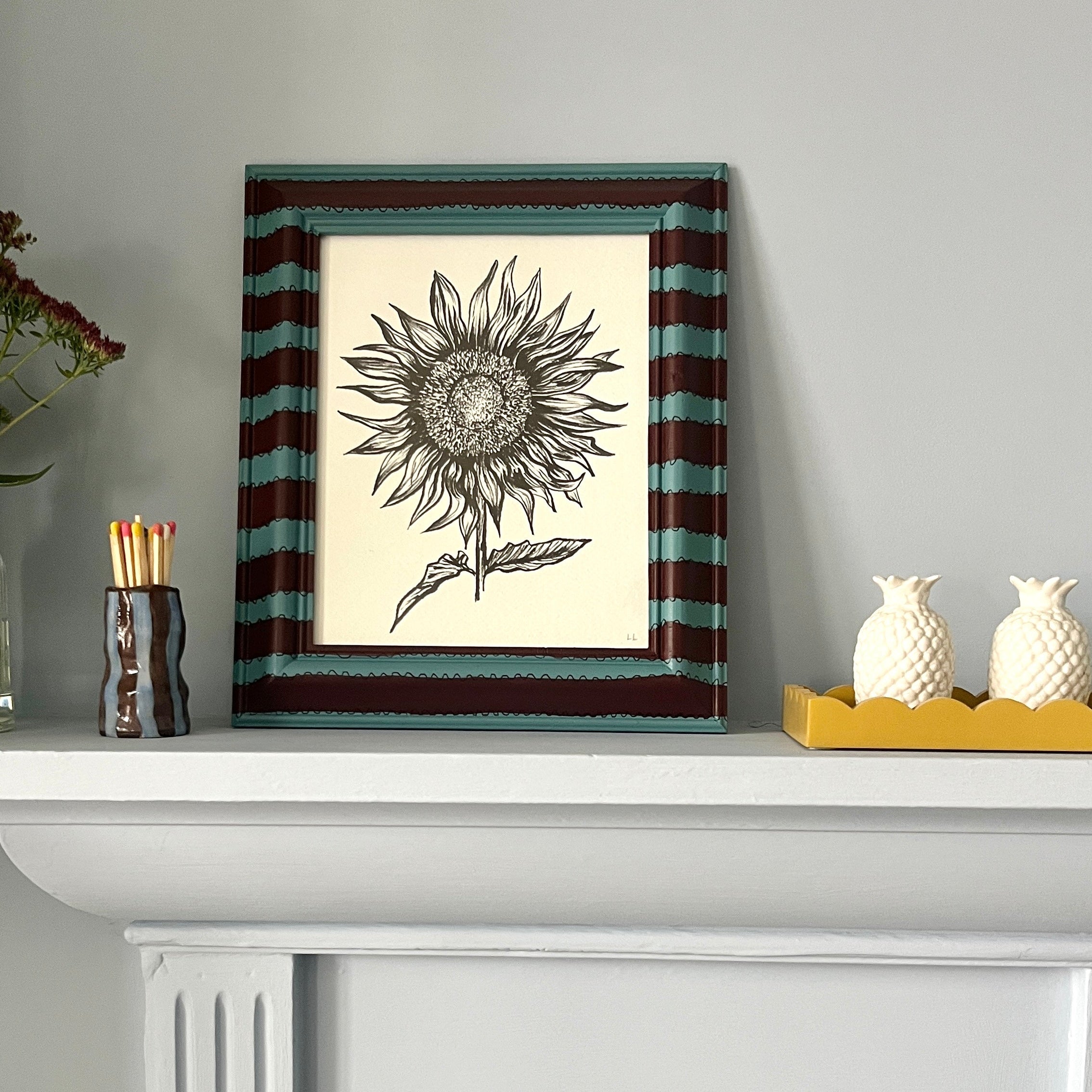 Framed artwork of a sunflower on a white shelf with decorative items.