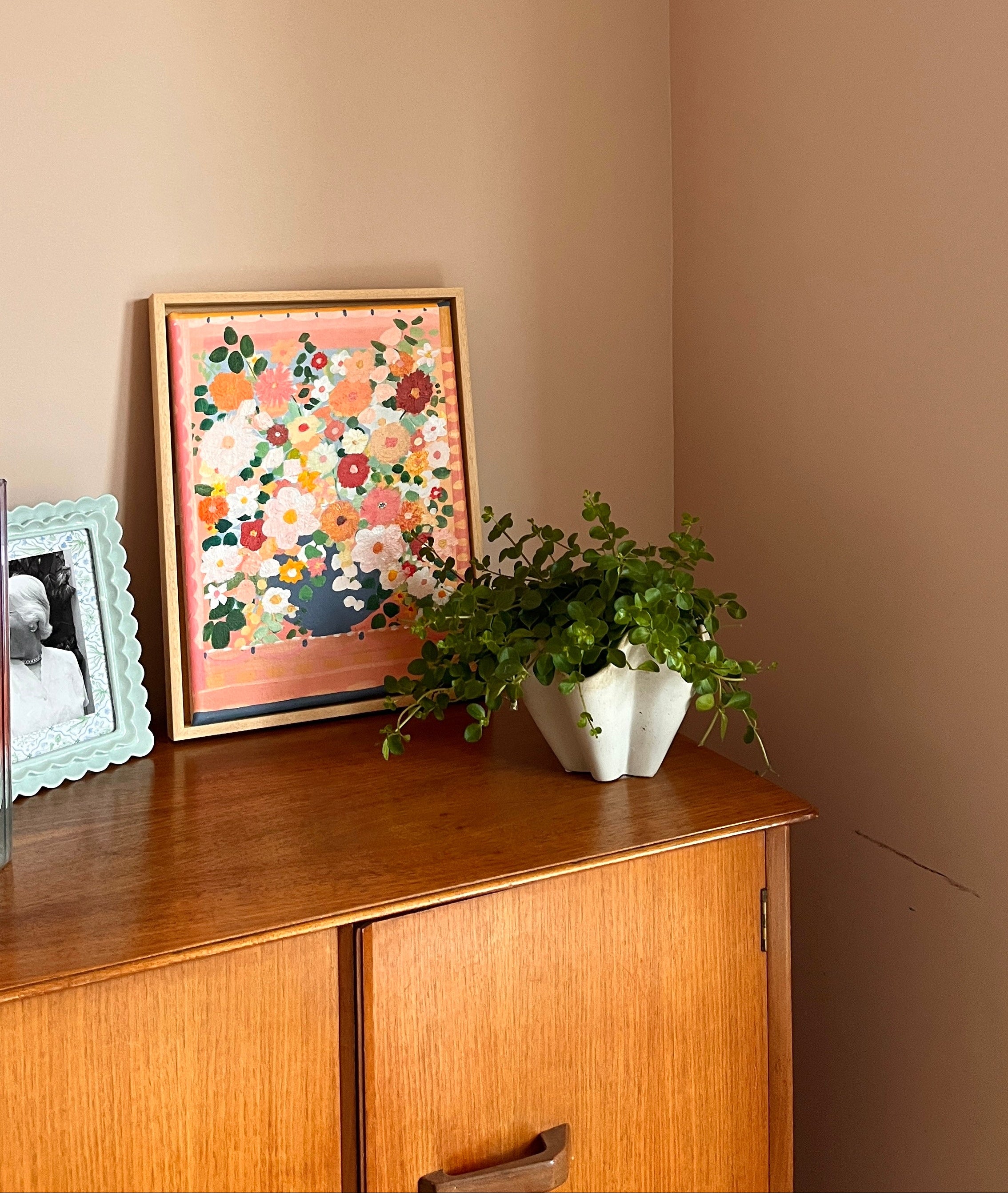 Framed floral artwork on a wooden sideboard against a beige wall with a wall-mounted light fixture.
