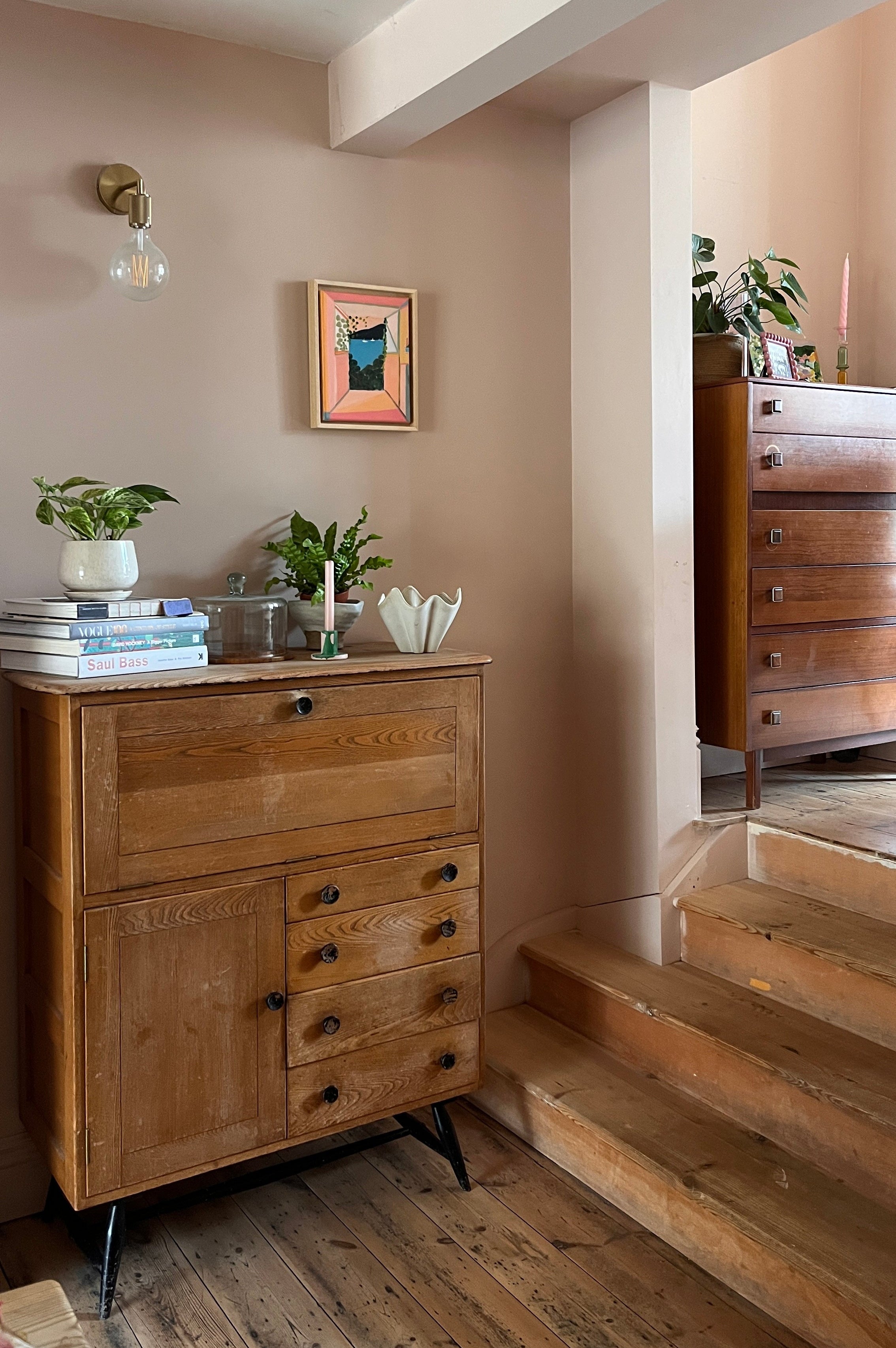 Wooden dresser with drawers in a room with a staircase and framed picture on the wall.