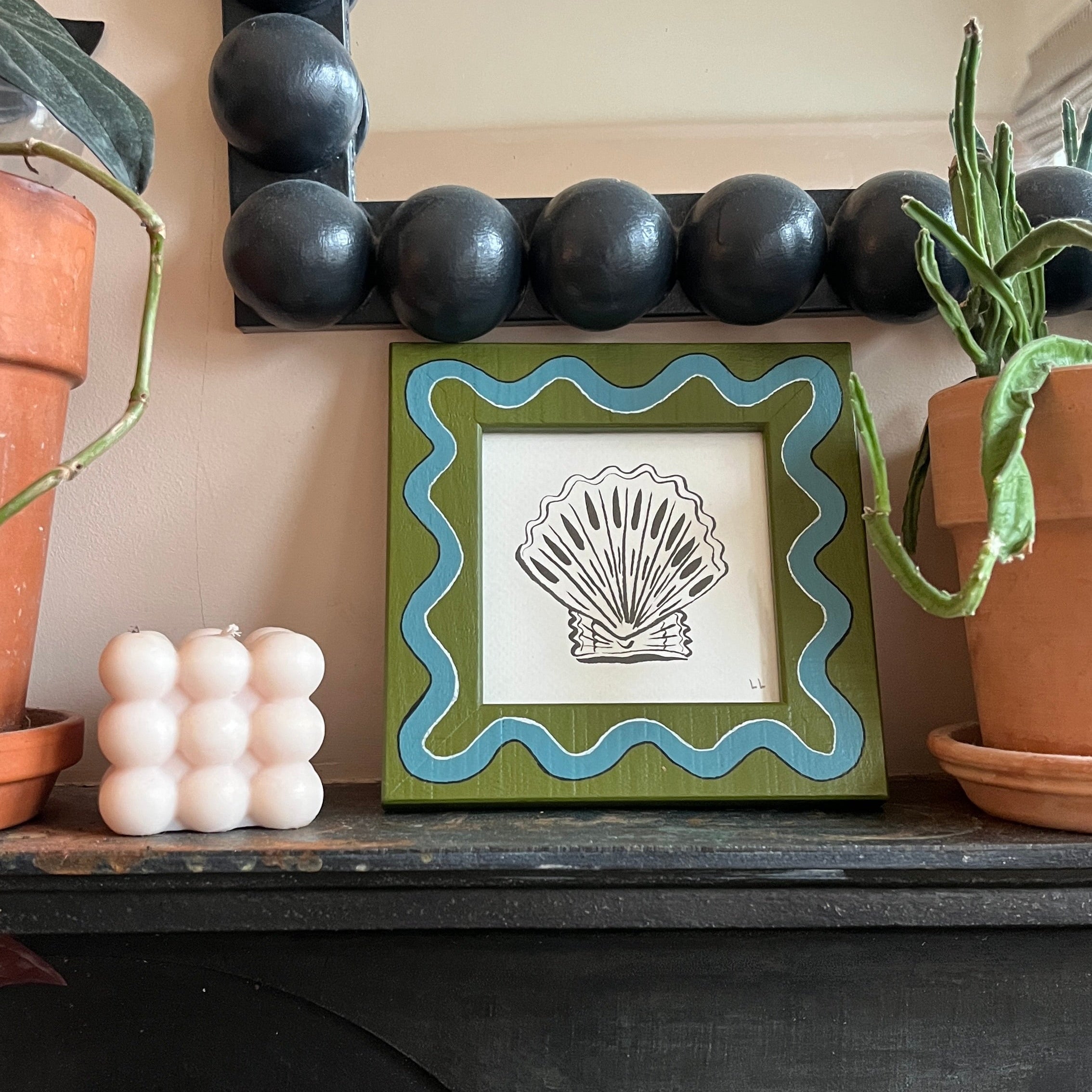 Decorative shelf with a framed artwork of a shell, surrounded by plants and decorative items.
