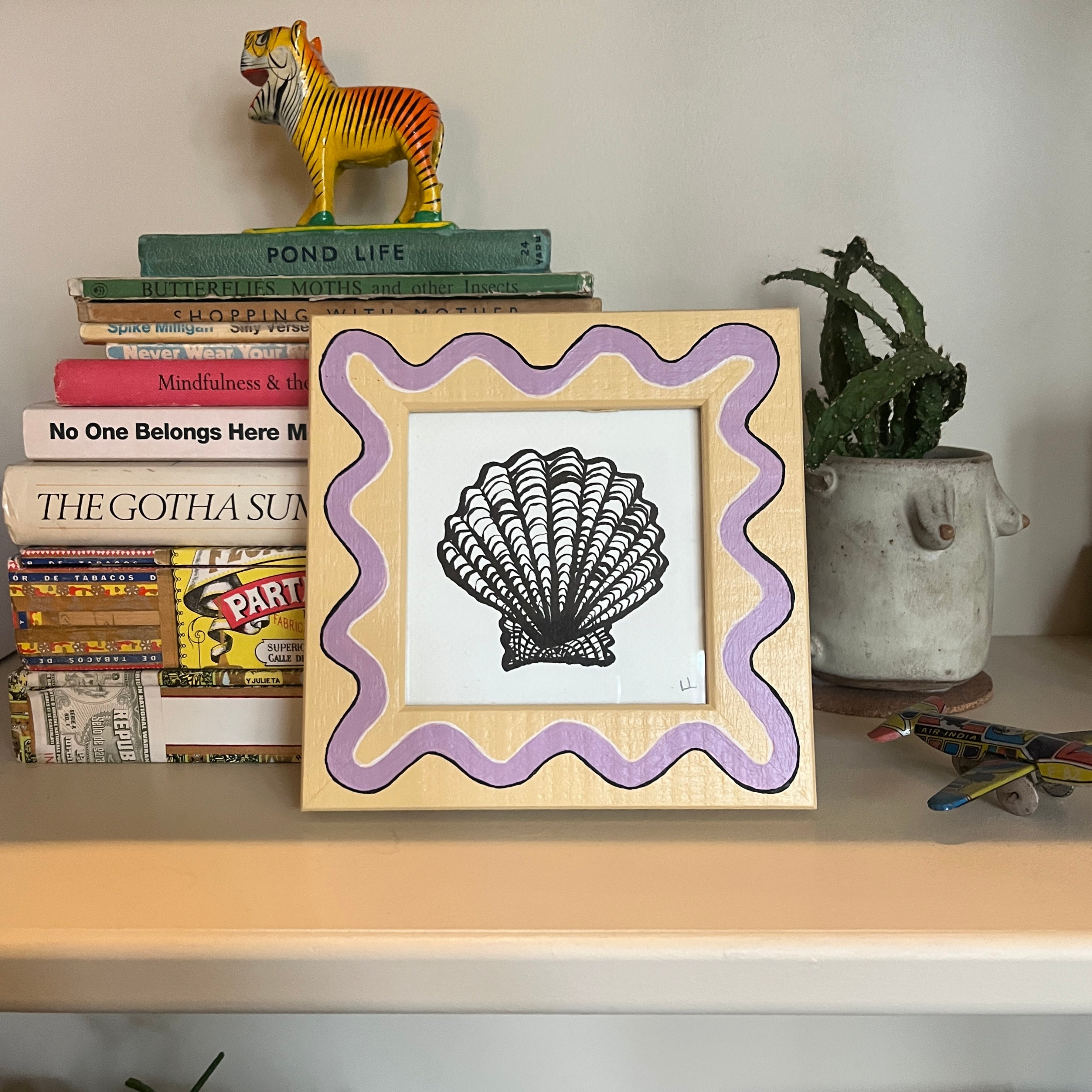 Framed artwork of a shell with a purple border on a shelf with books and a plant.