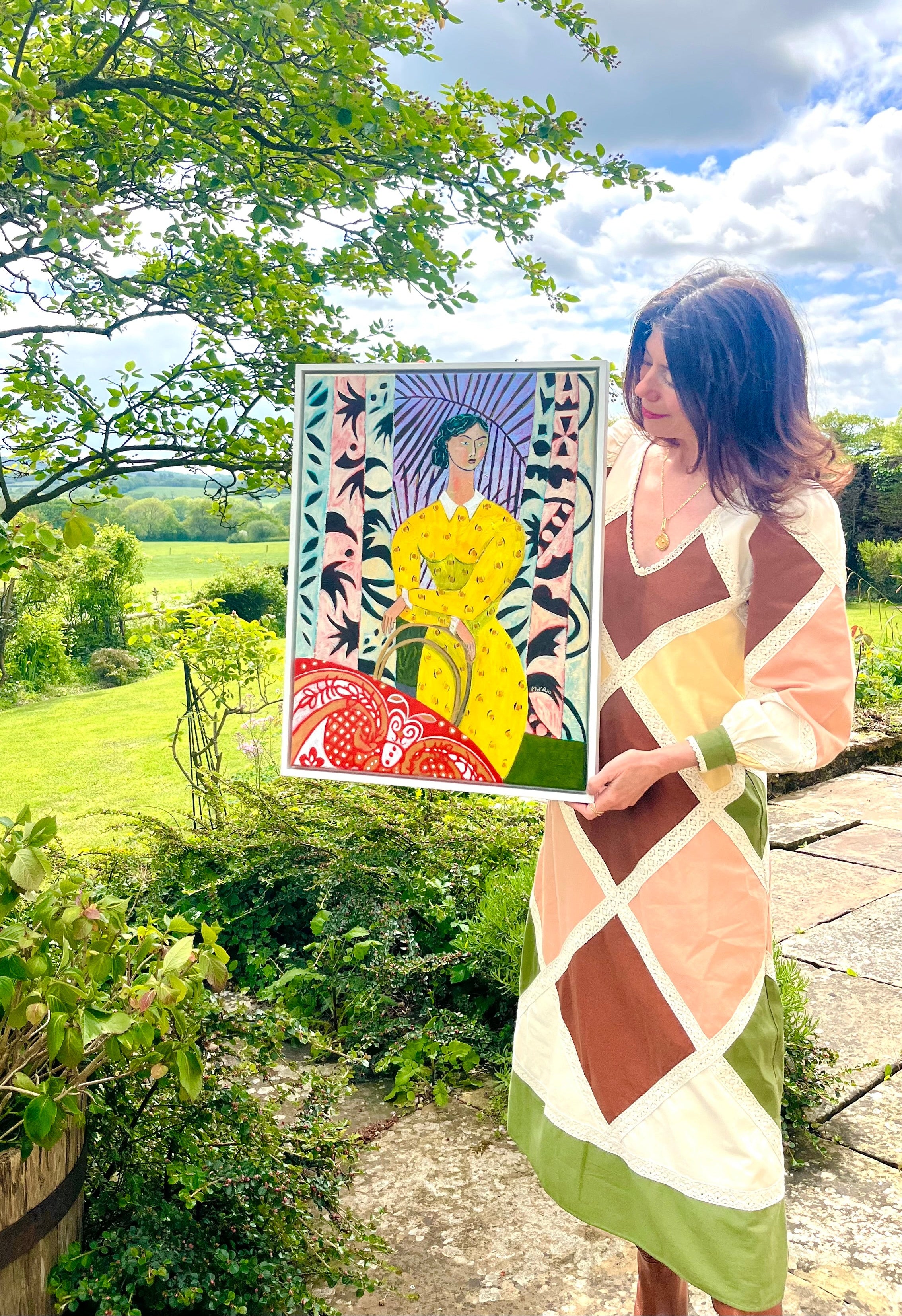 Woman holding a framed painting outdoors with greenery and blue sky in the background