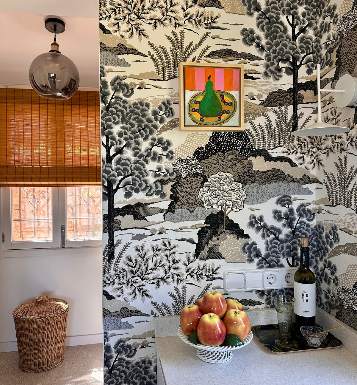 A kitchen interior featuring a bold black, white, and gray botanical and landscape wallpaper pattern. A white countertop holds a bowl of red apples, a wine bottle, and a glass, with a small framed colorful painting mounted on the wall.